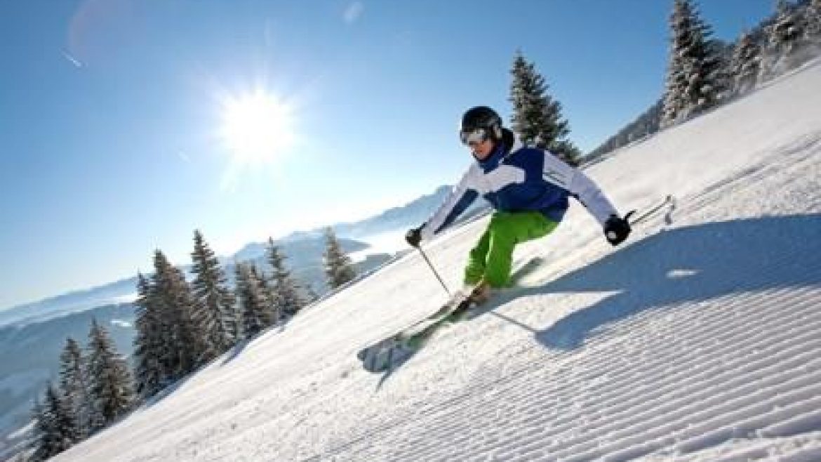 Person beim Skifahren auf einer sonnigen, verschneiten Piste mit Bäumen und Bergen im Hintergrund.