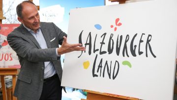 Ein Mann im Anzug gestikuliert in Richtung eines Schildes mit der Aufschrift Salzburger Land und bunten Formen um den Text herum.