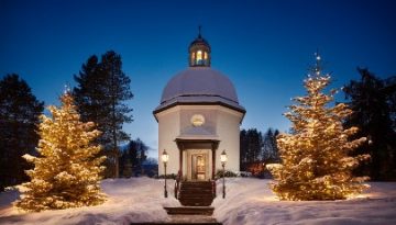 Eine schneebedeckte Kapelle mit beleuchteten Weihnachtsbäumen und Lampen an einem Winterabend.