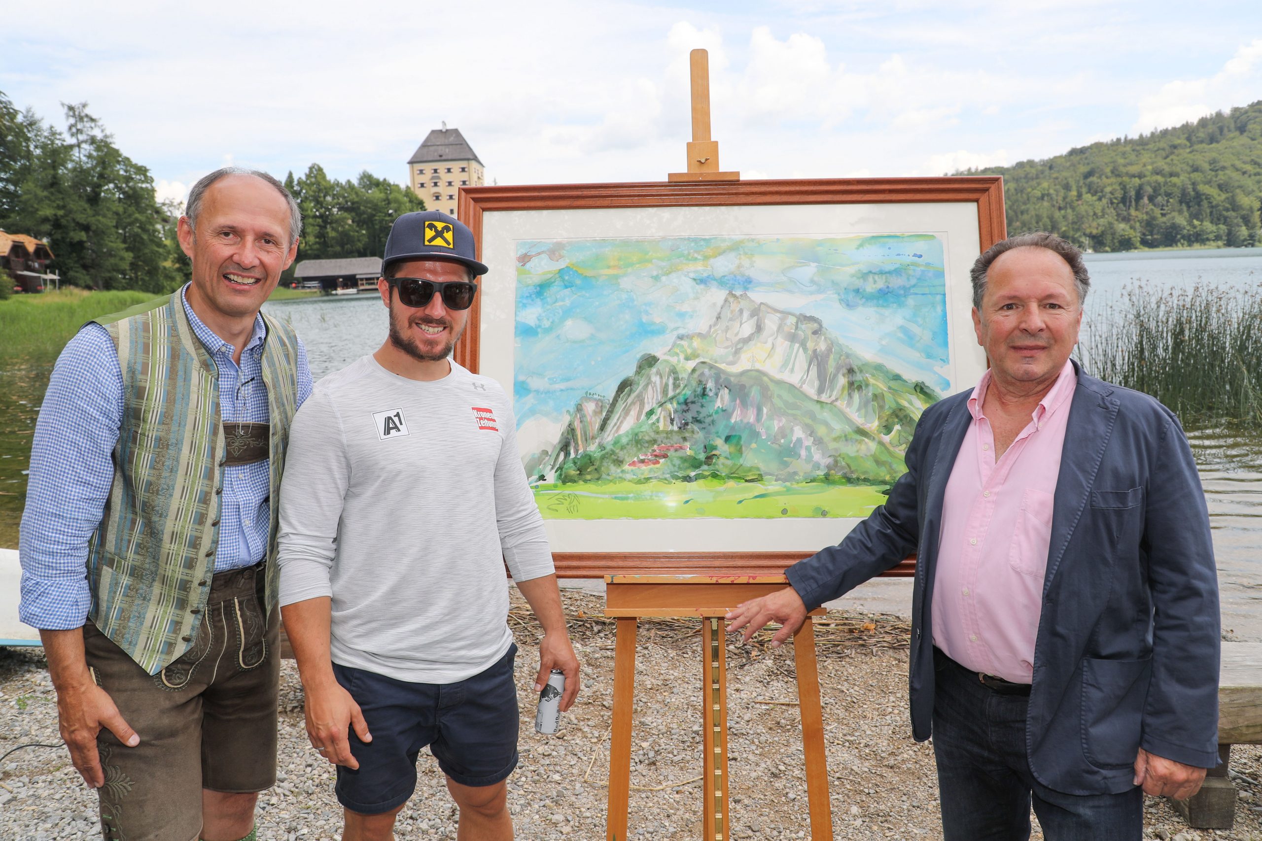 Drei Männer stehen an einem See, zwischen ihnen auf einer Staffelei ein buntes Berggemälde, inspiriert von Marcel Hirscher.