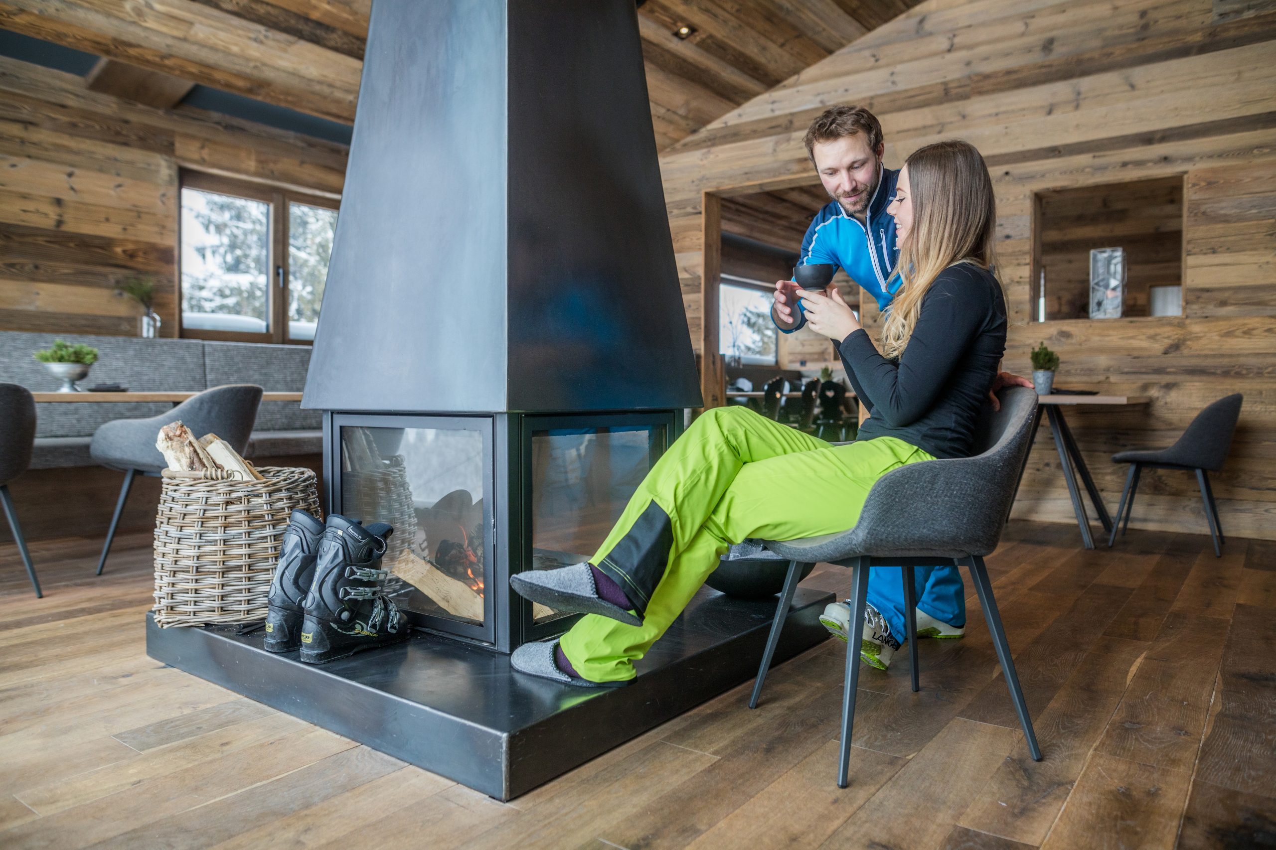 Ein Paar in Skikleidung sitzt an einem modernen Kamin und genießt heiße Getränke in einer gemütlichen CEE-Berghütte.
