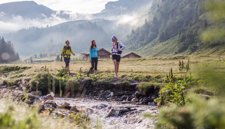 Drei Personen wandern entlang eines Baches in einem malerischen Bergtal mit Nebel und grünen Hügeln im Hintergrund.