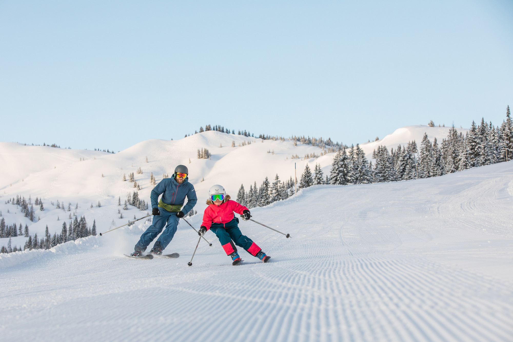 Ein Erwachsener und ein Kind fahren auf einer präparierten, verschneiten Piste mit Kiefern und Bergen im Hintergrund.