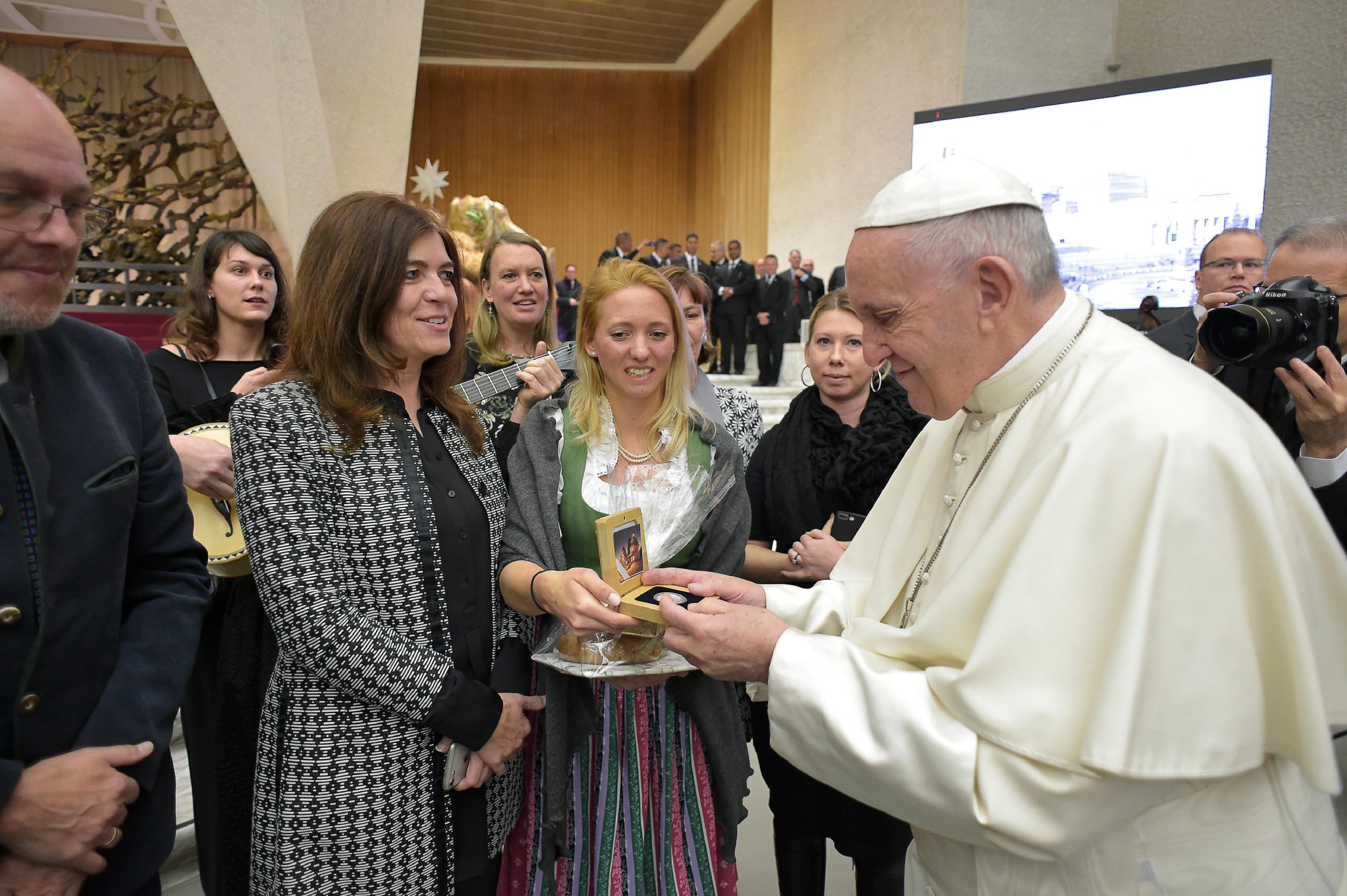 Der Papst begrüßt und tauscht Geschenke mit lächelnden Frauen in traditioneller Kleidung bei einer Veranstaltung in einer Halle aus.