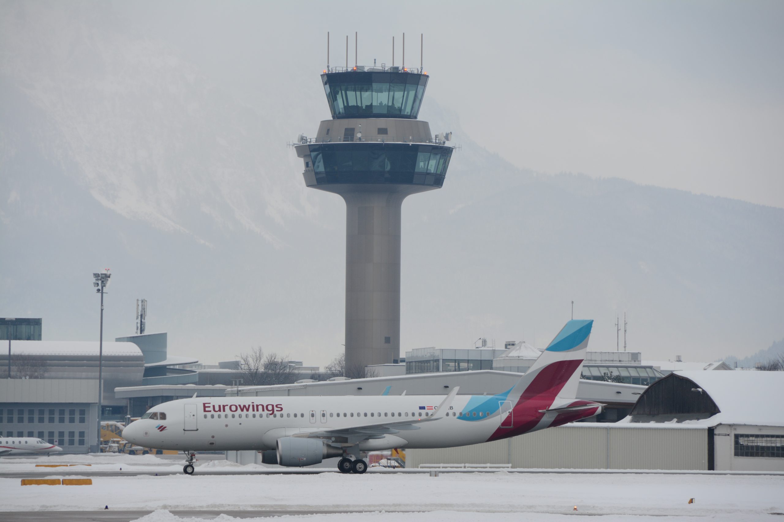 Ein Eurowings-Flugzeug rollt auf einer verschneiten Landebahn mit einem Flughafenkontrollturm im Hintergrund.