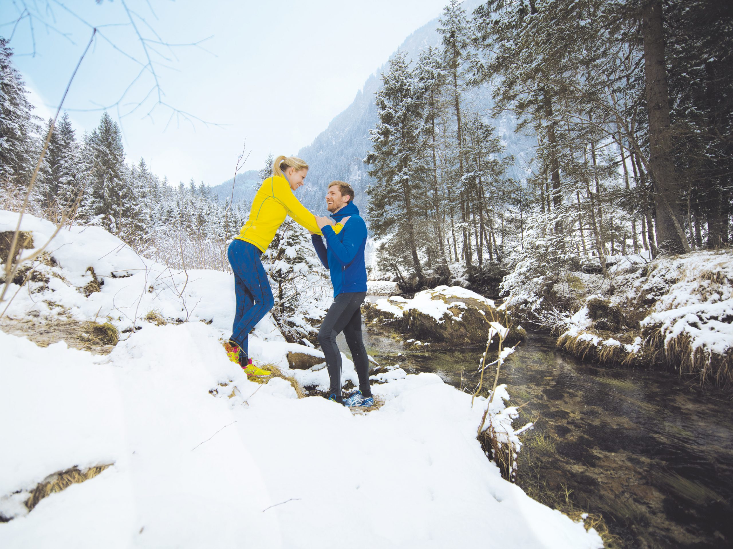 Zwei Personen in hellen Jacken stehen auf verschneitem Boden an einem Bach in einem Winterwald und lächeln sich an.