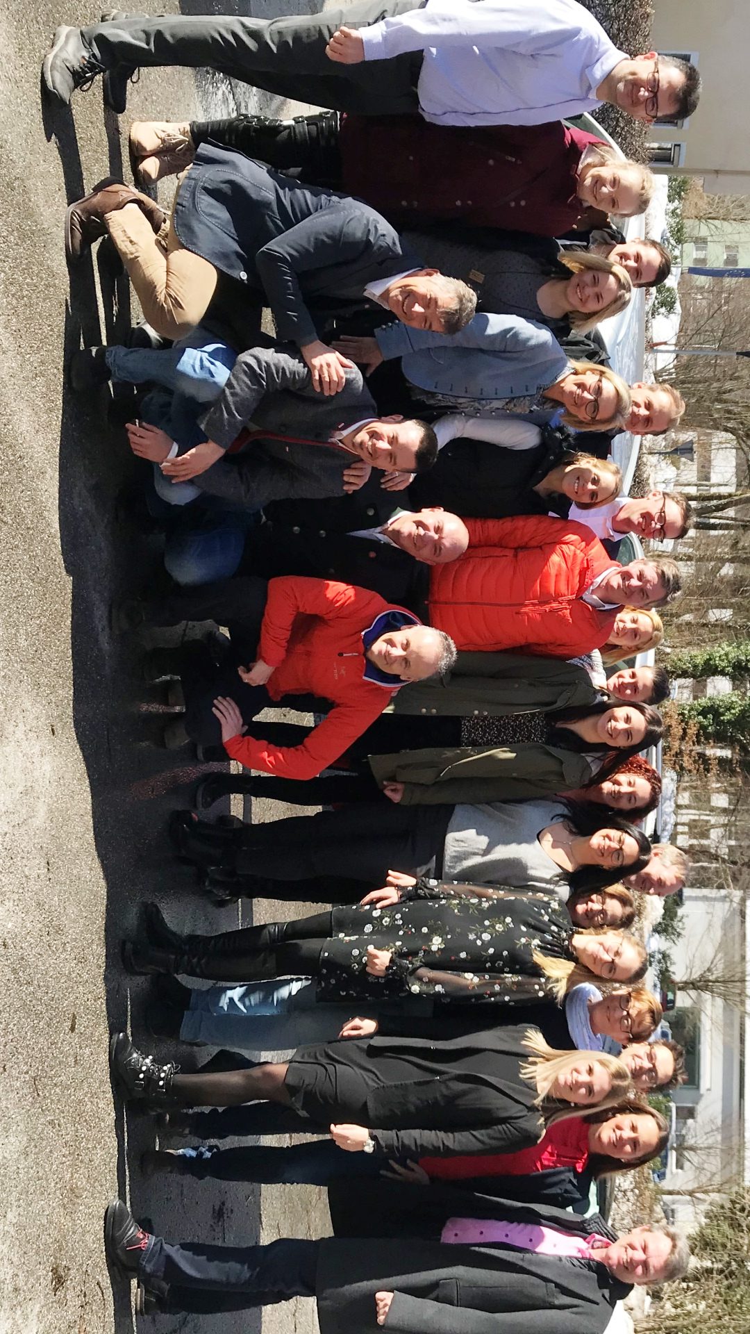 Eine Gruppe von Menschen posiert an einem sonnigen Tag im Freien, sie stehen und knien in Reihen und lächeln in die Kamera.
