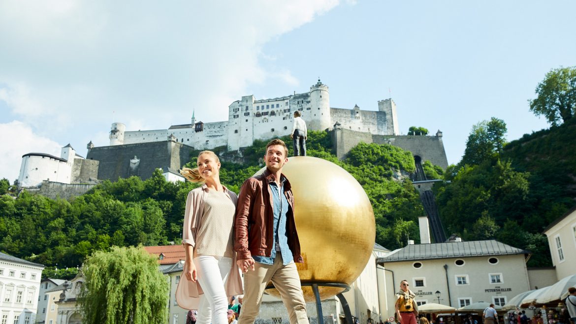 Ein lächelndes Paar spaziert über einen belebten Platz mit einer goldenen Kugelskulptur und einem Schloss auf einem Hügel im Hintergrund.