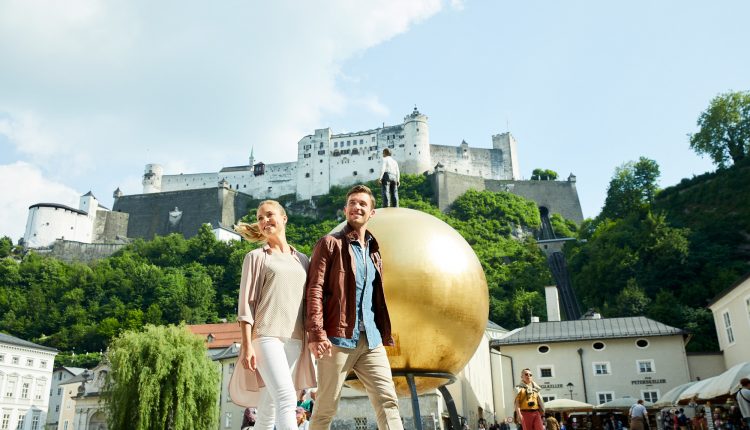 Ein lächelndes Paar spaziert über einen belebten Platz mit einer goldenen Kugelskulptur und einem Schloss auf einem Hügel im Hintergrund.
