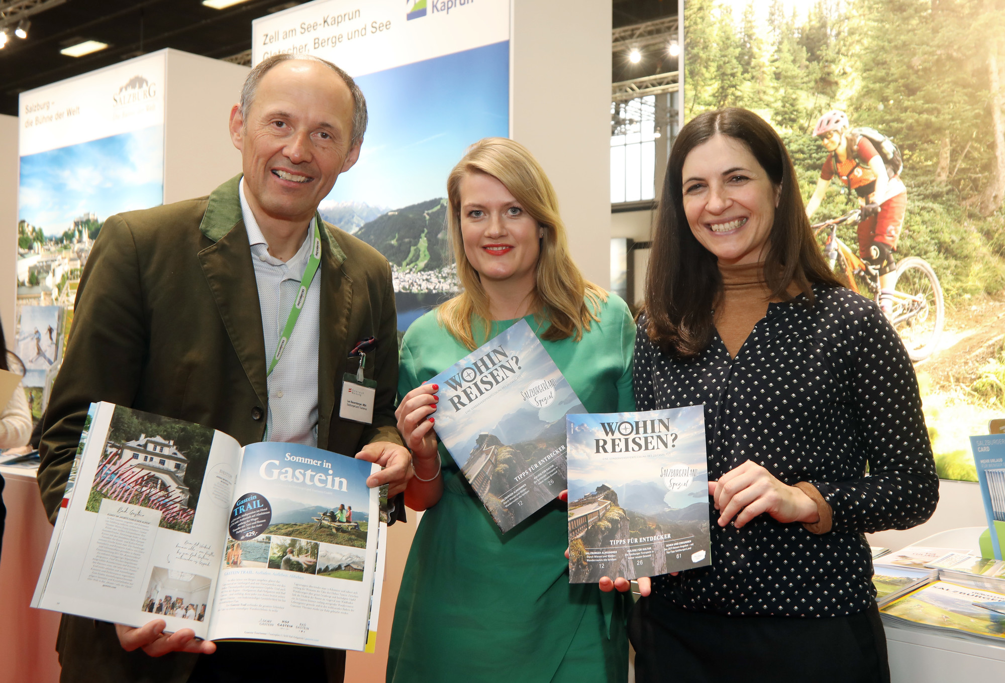 Drei Personen halten an einem Stand auf einer Tourismusmesse Reisemagazine und Broschüren hoch, im Hintergrund sind Bilder von Bergen zu sehen.