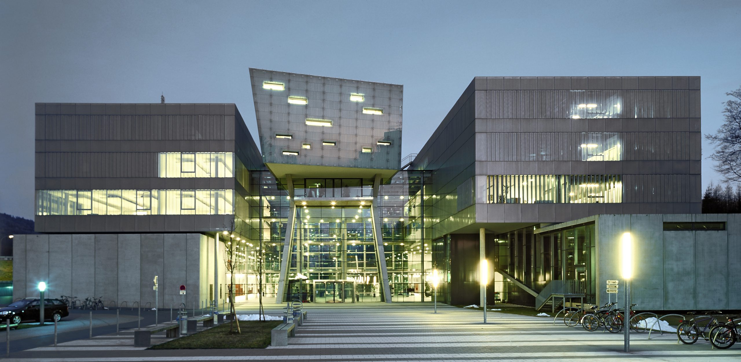 Modernes Gebäude aus Glas und Beton mit geometrischer Architektur, das in der Abenddämmerung beleuchtet wird, vor dem Fahrräder geparkt sind.