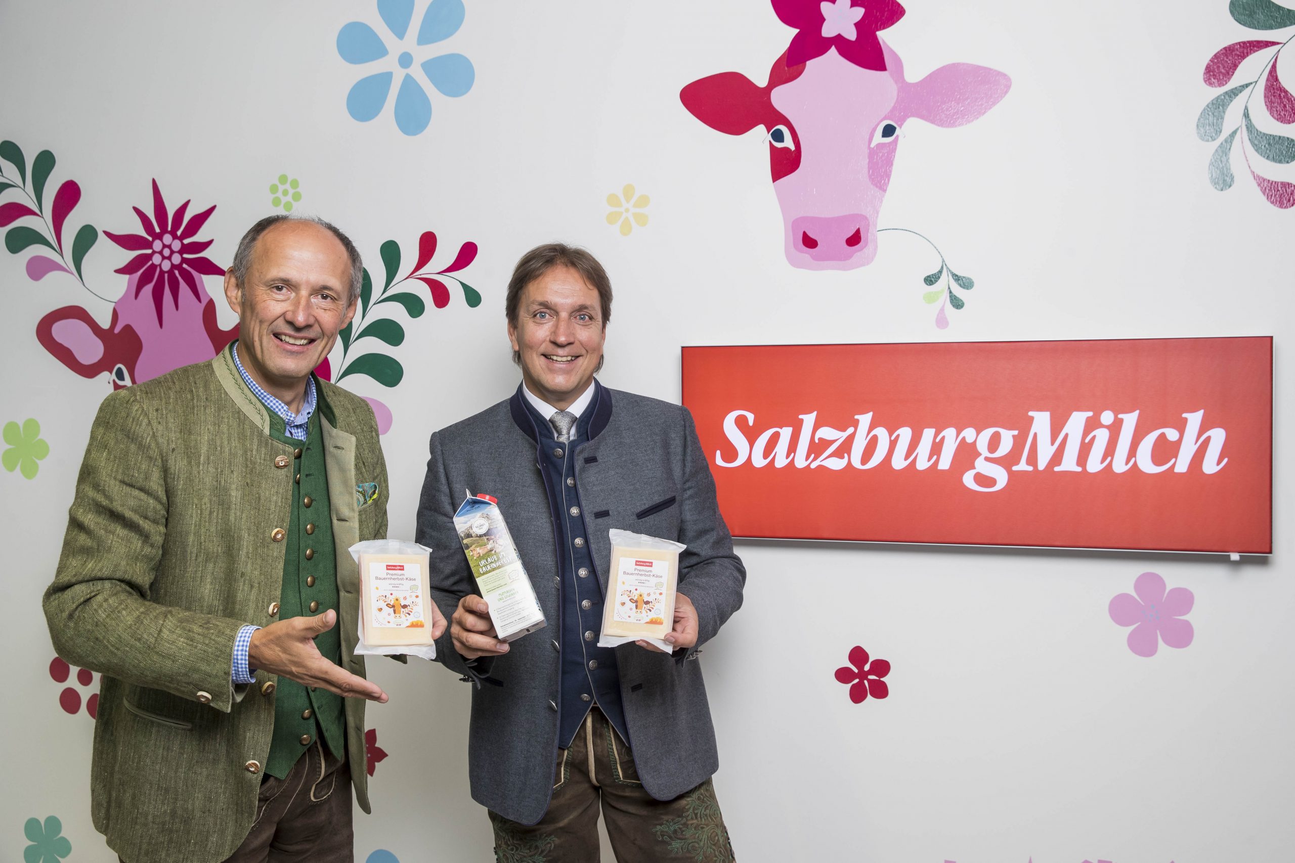 Zwei Männer in traditioneller österreichischer Kleidung halten SalzburgMilch-Milchprodukte in der Hand und stehen neben einem roten SalzburgMilch-Schild.