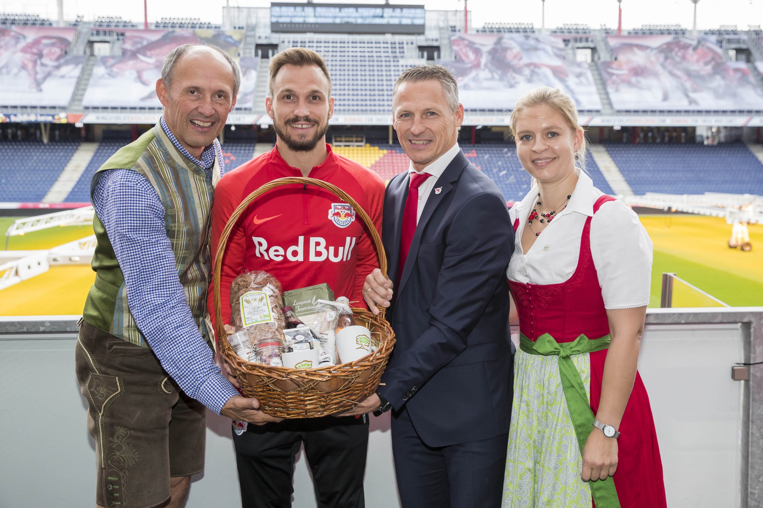 Vier lächelnde Menschen mit einem Geschenkkorb in einem Stadion, zwei in traditioneller Kleidung und zwei in moderner Kleidung.