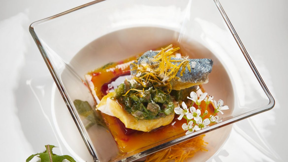Feinschmecker-Fischgericht mit Sauce, Kräutern und essbaren Blüten, serviert in einer quadratischen Glasschale auf einem weißen Teller.