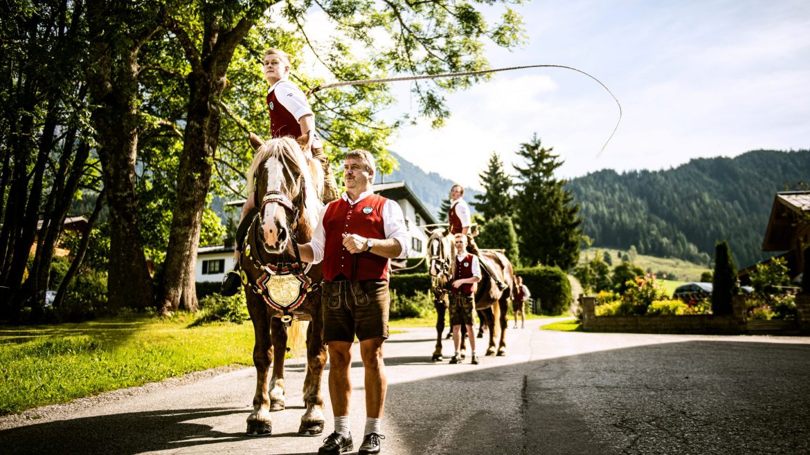 Männer in österreichischer Tracht führen Pferde mit Reitern über eine sonnige Landstraße, umgeben von Bäumen und Bergen.