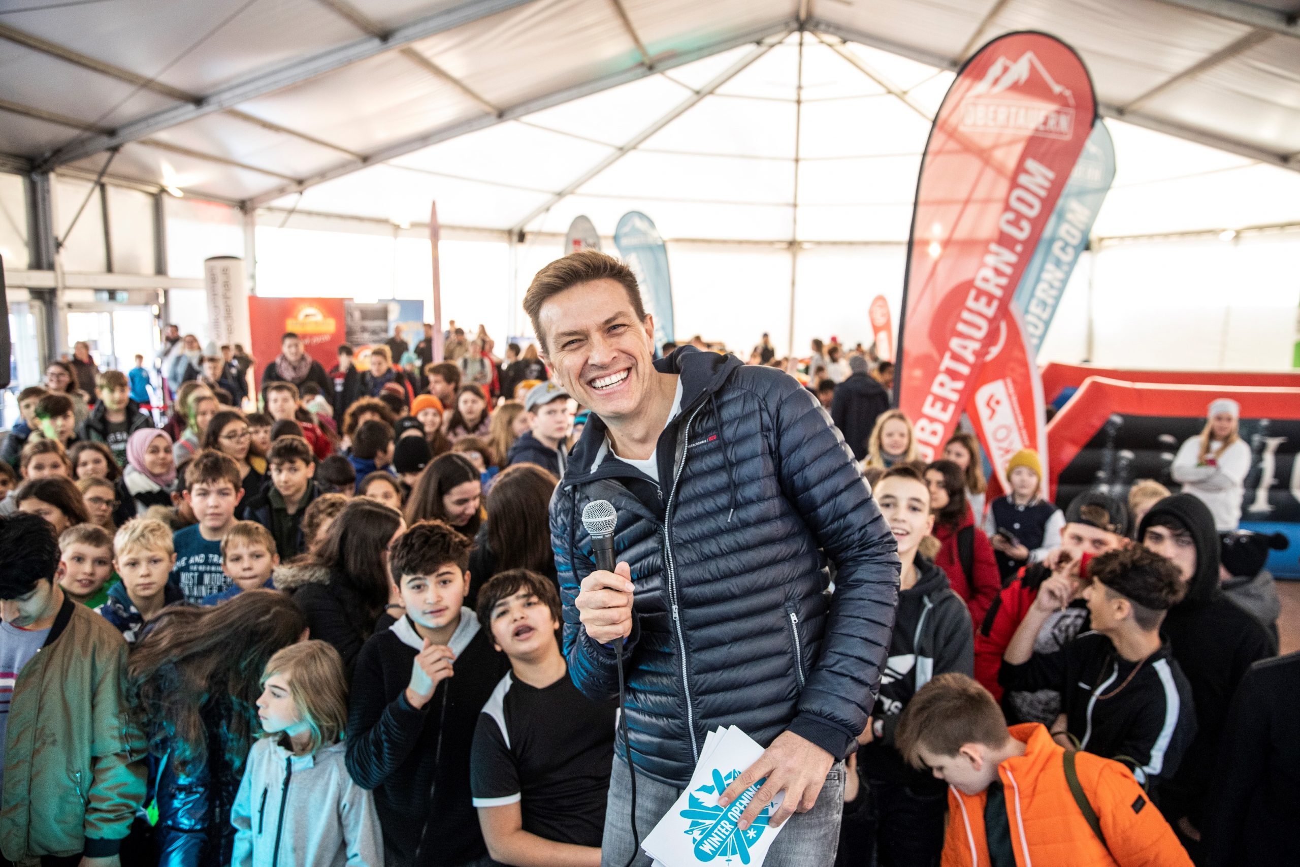 Ein Mann mit einem Mikrofon lächelt bei einer überfüllten Veranstaltung in einer Halle mit vielen Kindern und bunten Bannern.
