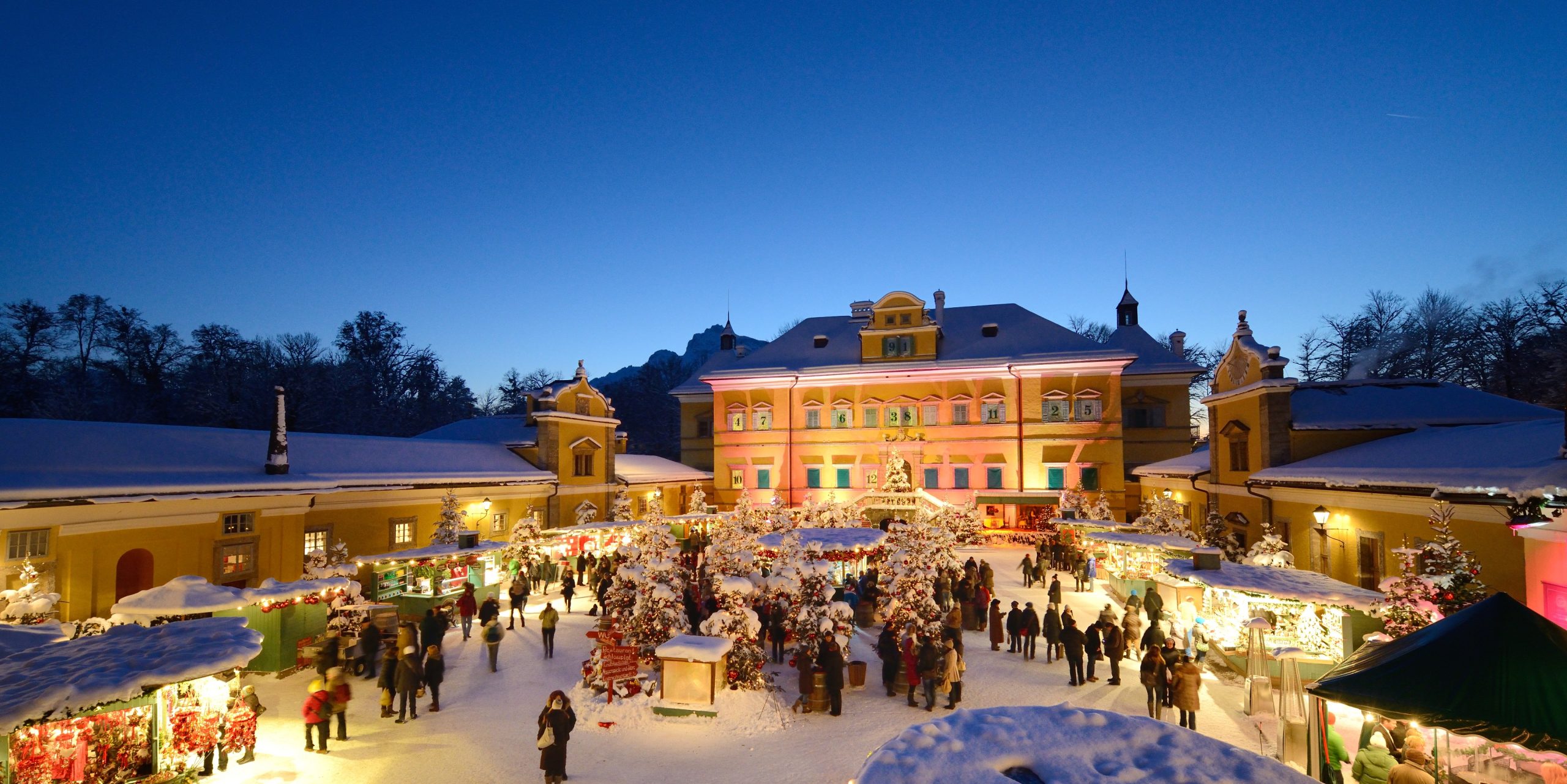Ein festlicher Weihnachtsmarkt bei Nacht mit bunten Lichtern, geschmückten Ständen und Menschen, die im Schnee spazieren gehen.