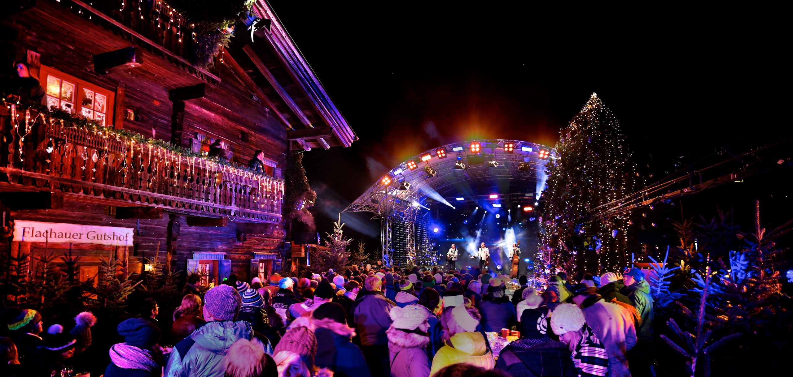Eine festliche Menschenmenge genießt ein lebhaftes nächtliches Konzert im Freien, umgeben von Lichtern und geschmückten Gebäuden.
