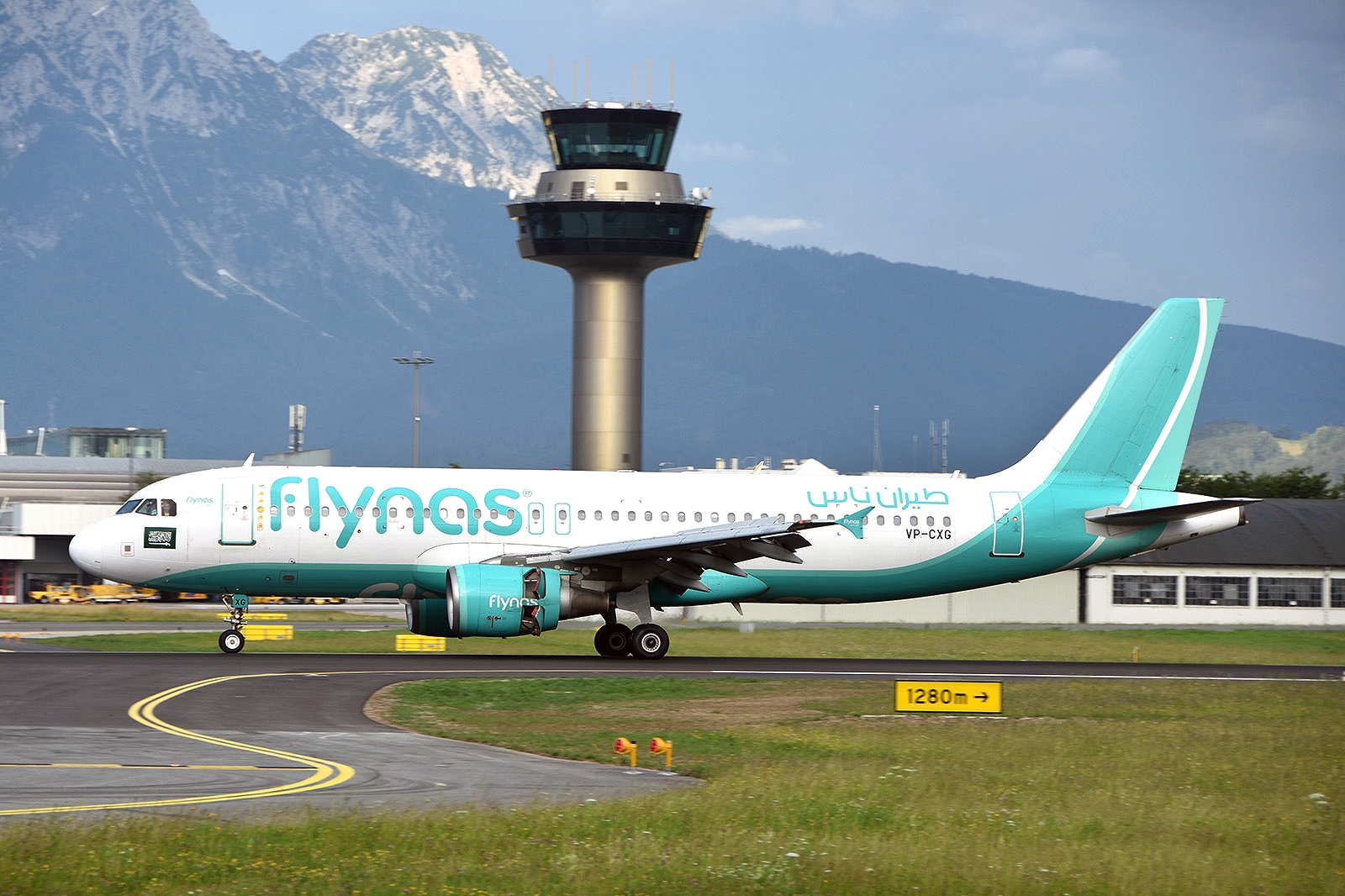 Ein Flynas-Flugzeug rollt auf einer Landebahn mit einem Kontrollturm und Bergen im Hintergrund.