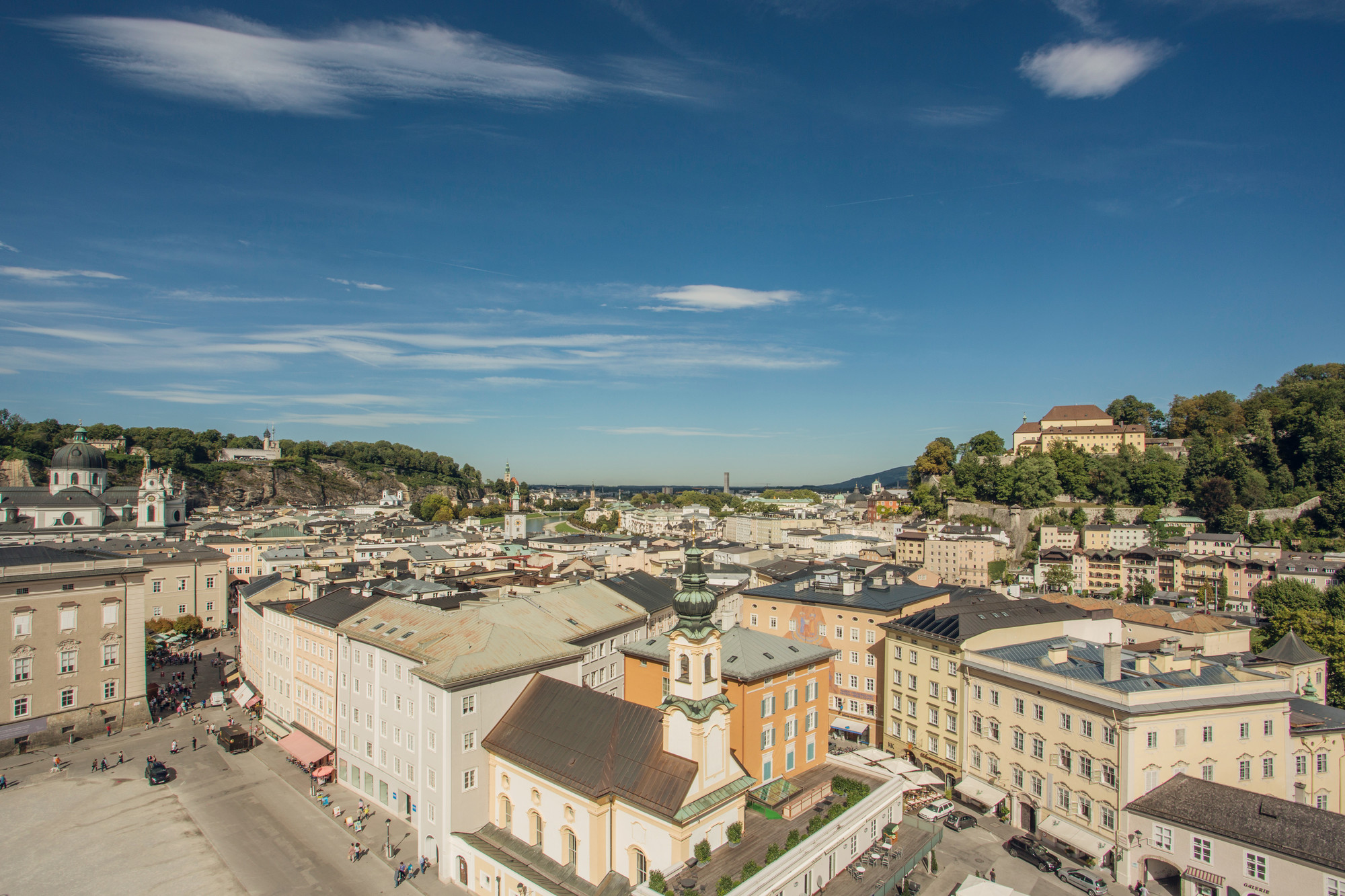 Luftaufnahme von Salzburg, Österreich, mit farbenfrohen historischen Gebäuden und Hügeln unter einem klaren blauen Himmel.