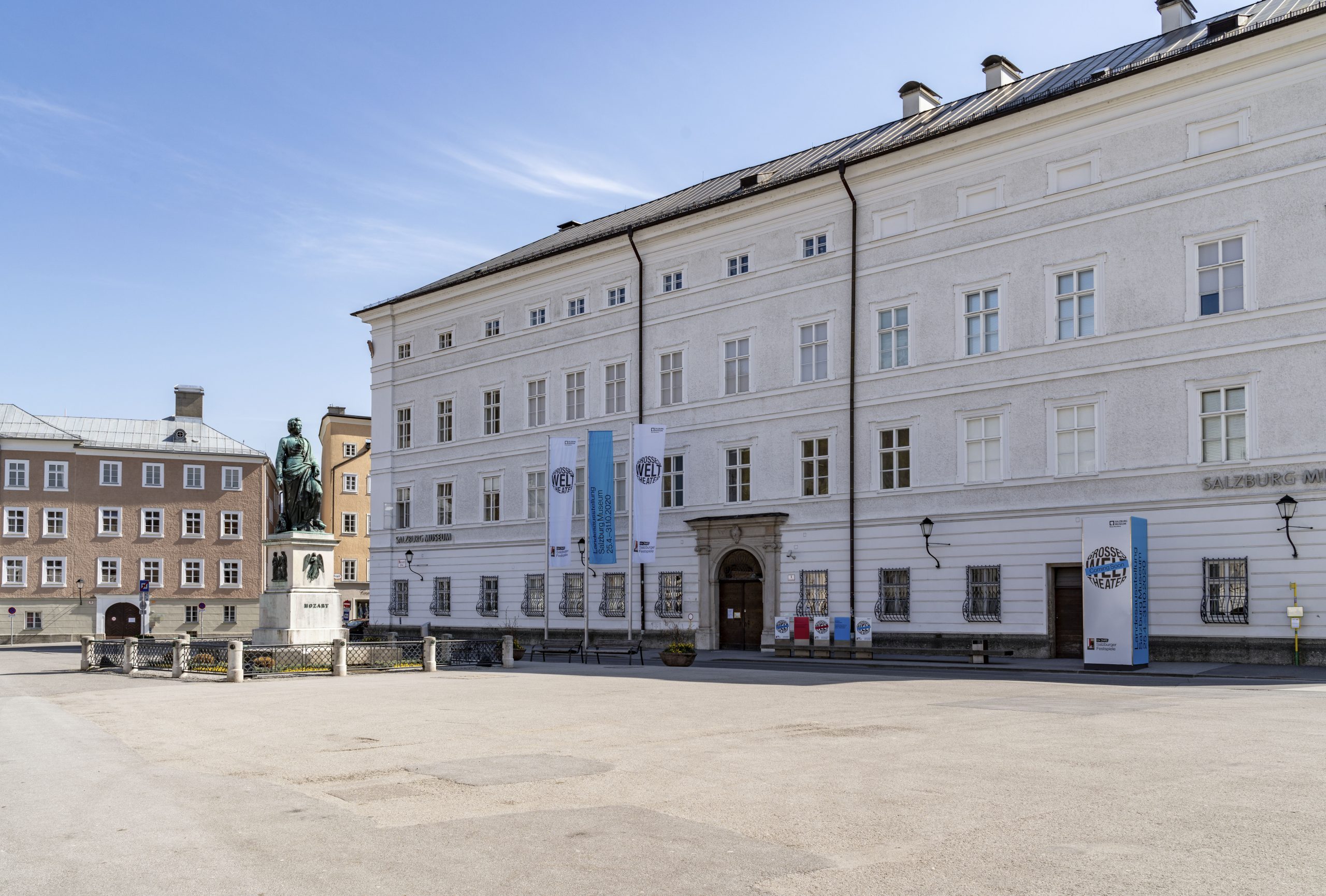 Großes weißes historisches Gebäude mit Fahnen und Statue auf einem ruhigen, offenen Platz an einem sonnigen Tag.