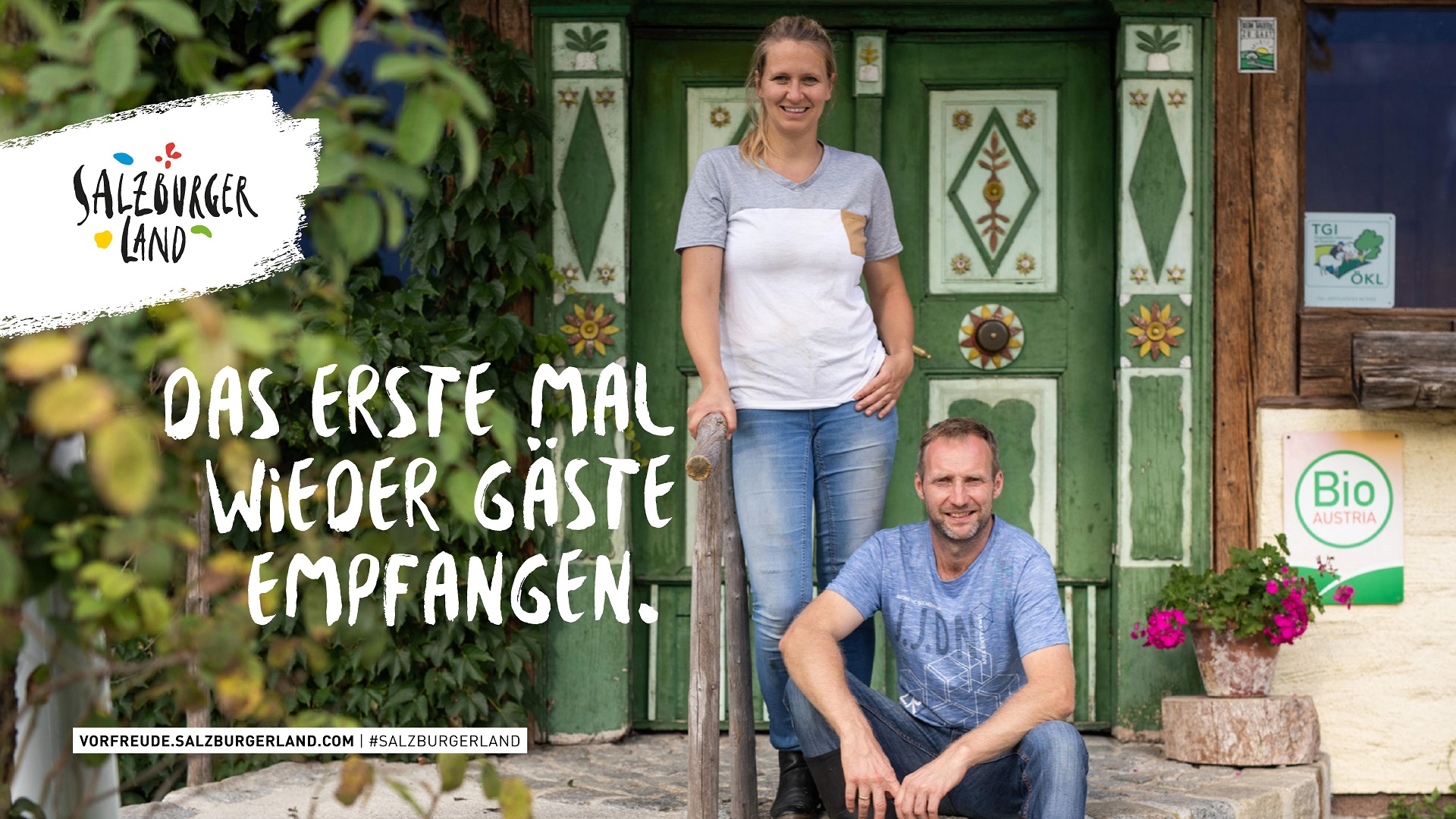 Ein lächelnder Mann und eine lächelnde Frau stehen vor einer rustikalen Holztür mit dem Text Das erste Mal wieder Gäste empfangen.