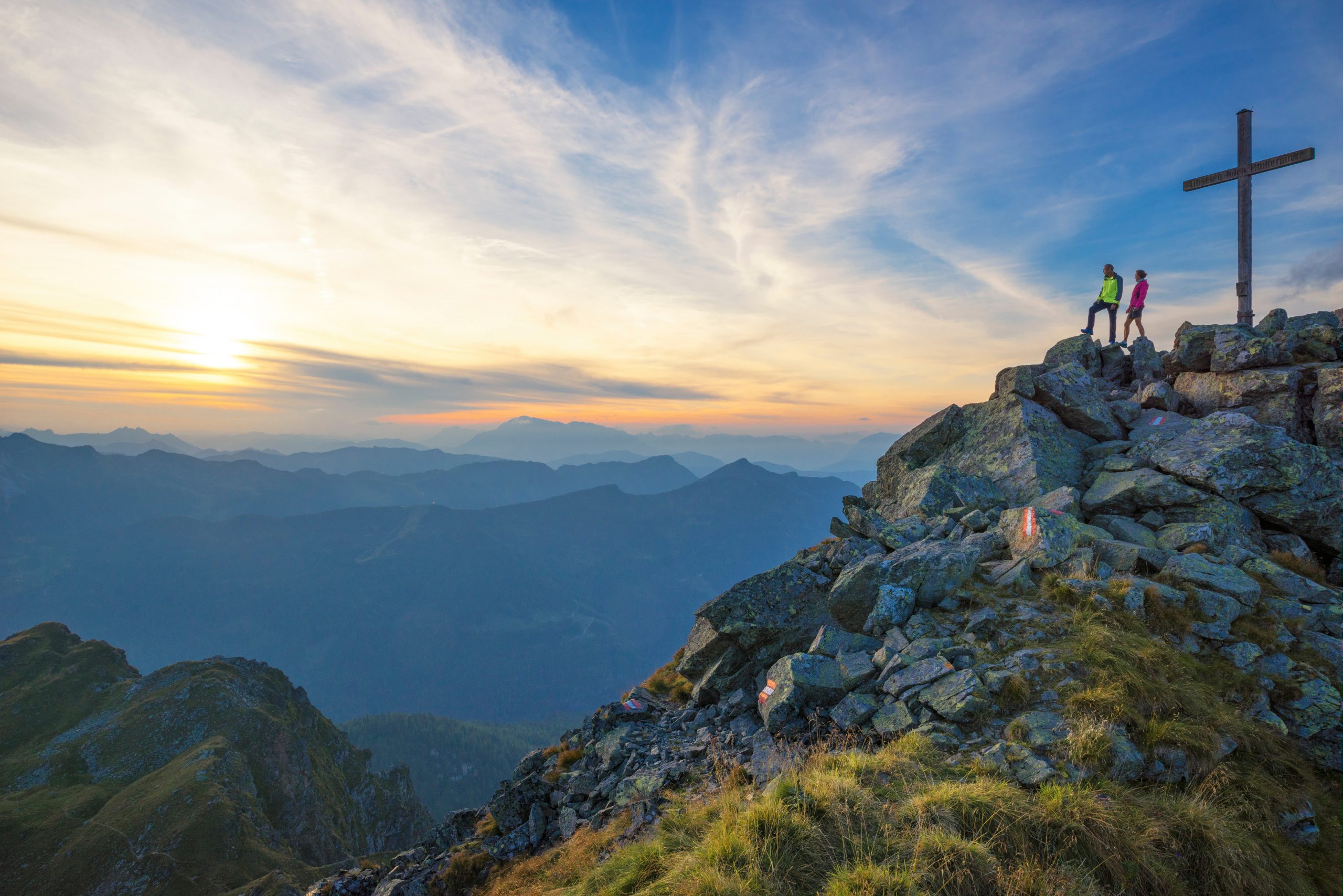 Zwei Wanderer stehen bei Sonnenuntergang an einem großen Kreuz auf einem felsigen Berggipfel und blicken auf ferne Hügel und Täler.