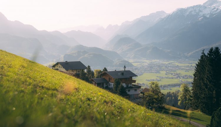 Häuser auf einem grasbewachsenen Hügel überblicken ein grünes Tal mit Bergen im Hintergrund unter einem dunstigen Himmel.