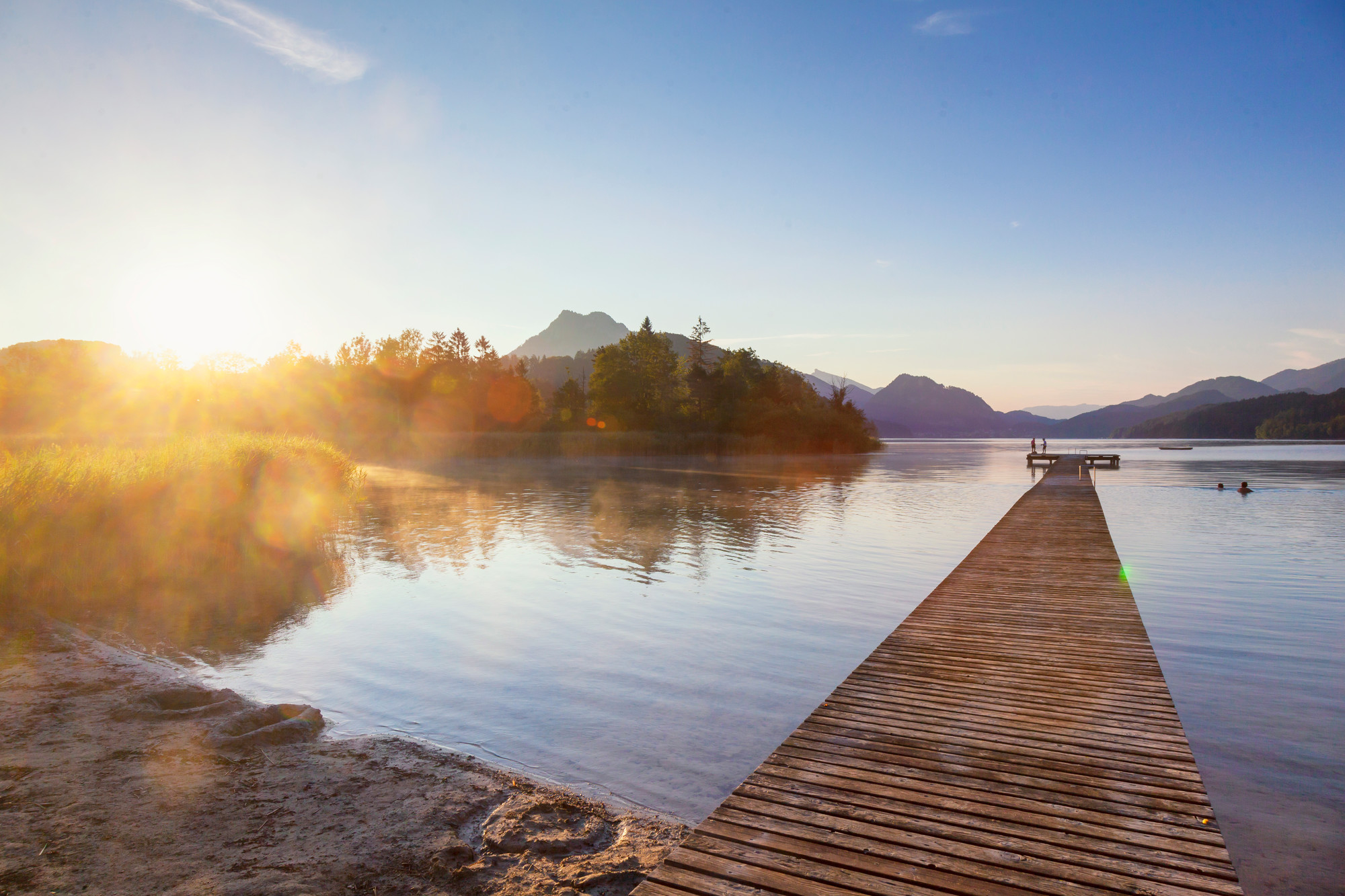 Der hölzerne Steg führt in einen ruhigen See bei Sonnenaufgang, mit Bäumen und Bergen in der Ferne.