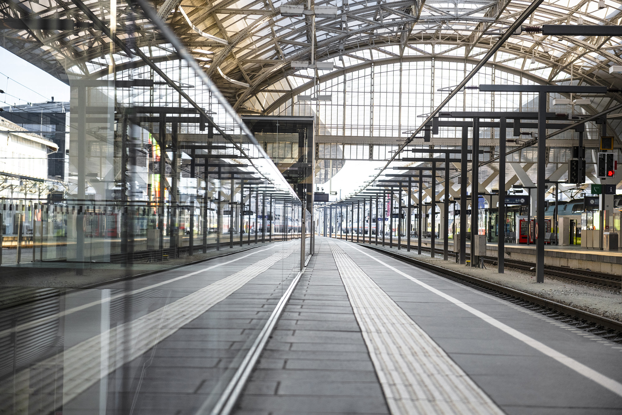 Leerer Bahnsteig mit Metallträgern, Glasdach und Gleisen, die sich in der Glaswand des Bahnhofs spiegeln.