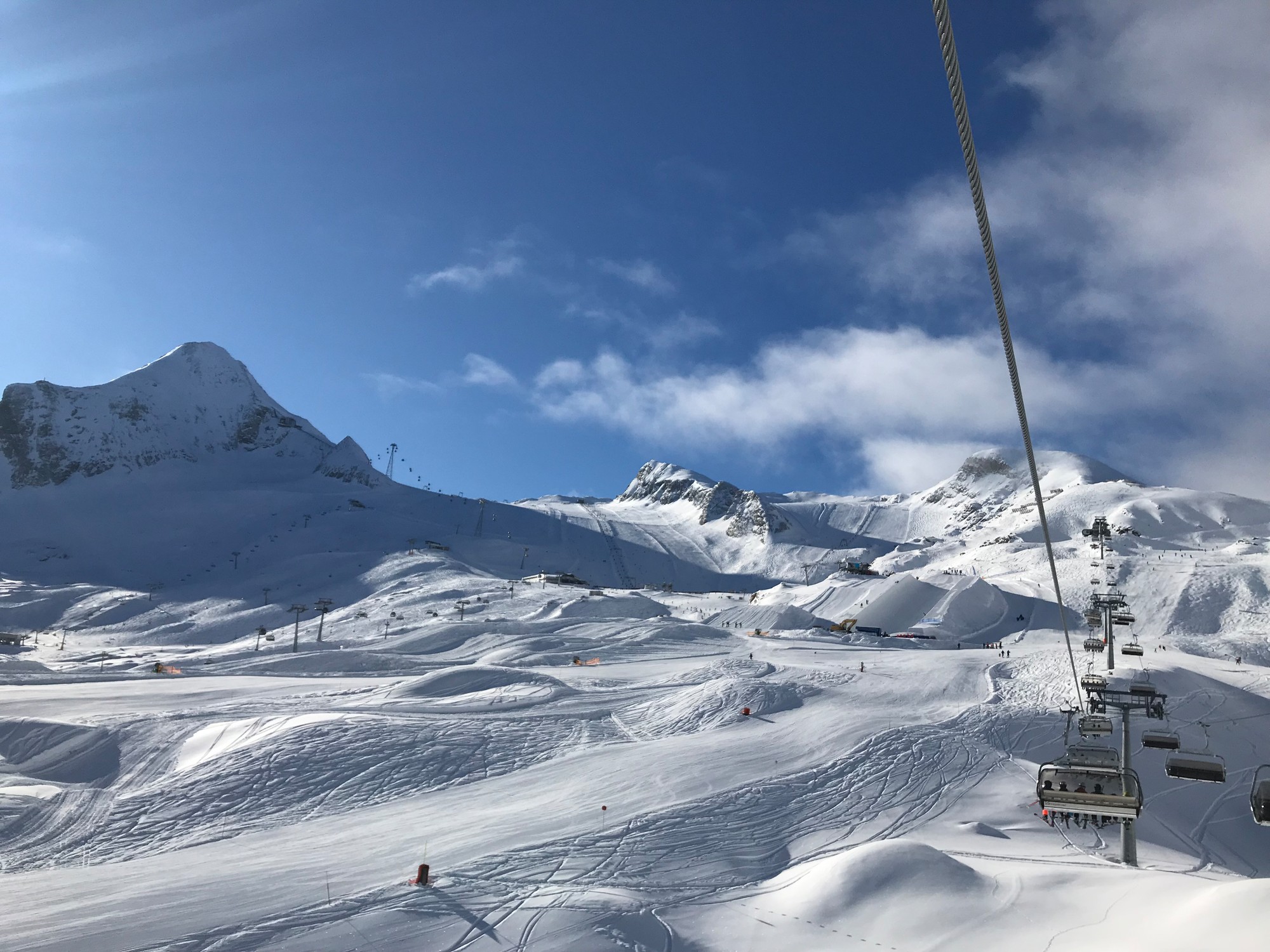 Skilift, der über verschneite Berge unter einem blauen Himmel mit Sonnenlicht und vereinzelten Wolken aufsteigt.