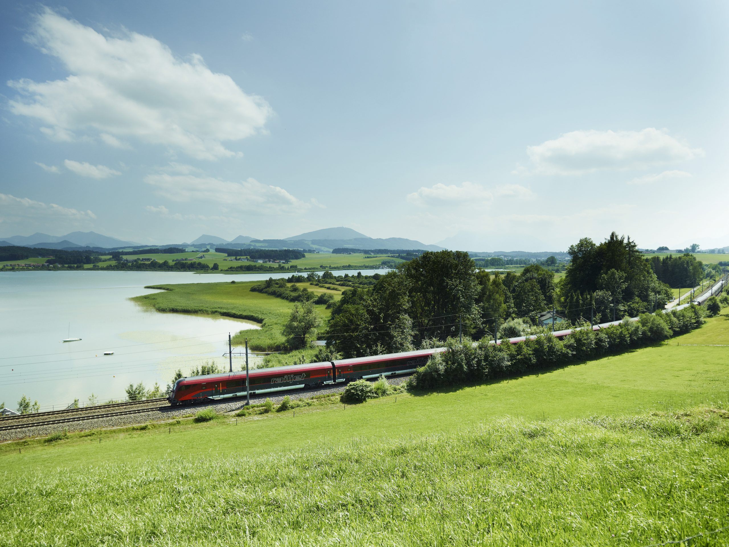 Ein roter Zug fährt an einem See entlang durch eine grüne Landschaft unter einem klaren blauen Himmel.