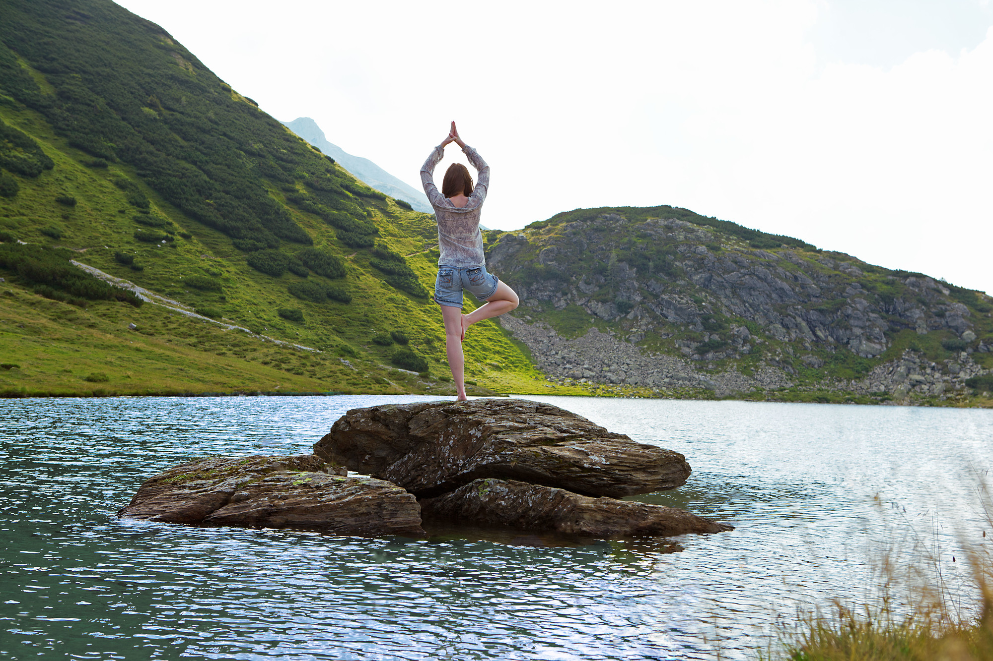 Person beim Yoga auf einem Felsen in einem See, umgeben von grünen Hügeln und hellem Tageslicht.