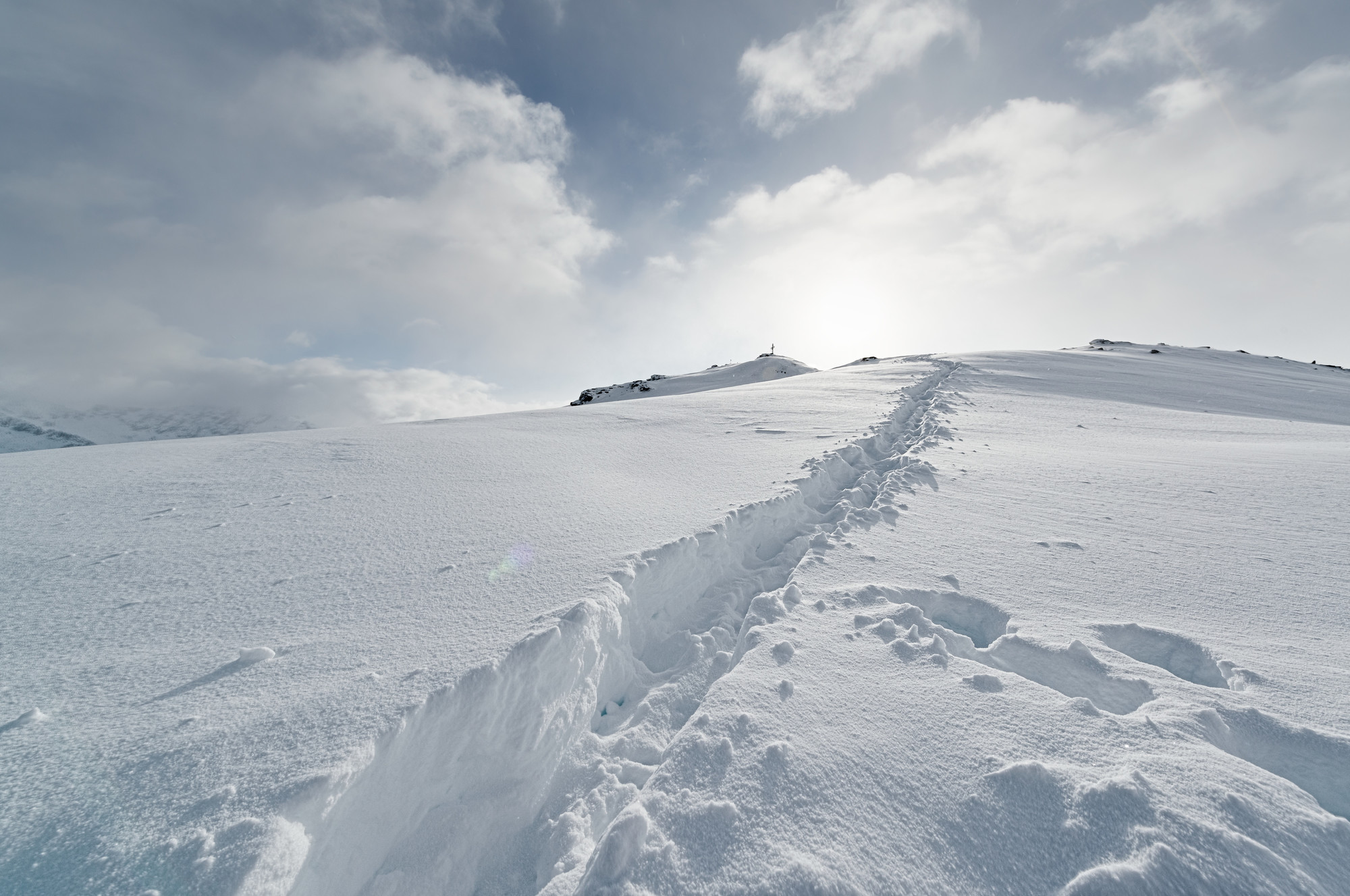 Ein schneebedeckter Weg mit Fußspuren führt unter einem wolkenverhangenen Himmel, durch den das Sonnenlicht bricht, einen sanften Hügel hinauf.