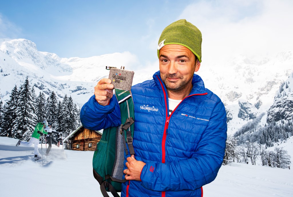 Ein Mann in blauer Jacke und grüner Mütze hält eine Snackbar in den verschneiten Bergen mit einer Hütte und Skifahrern im Hintergrund.