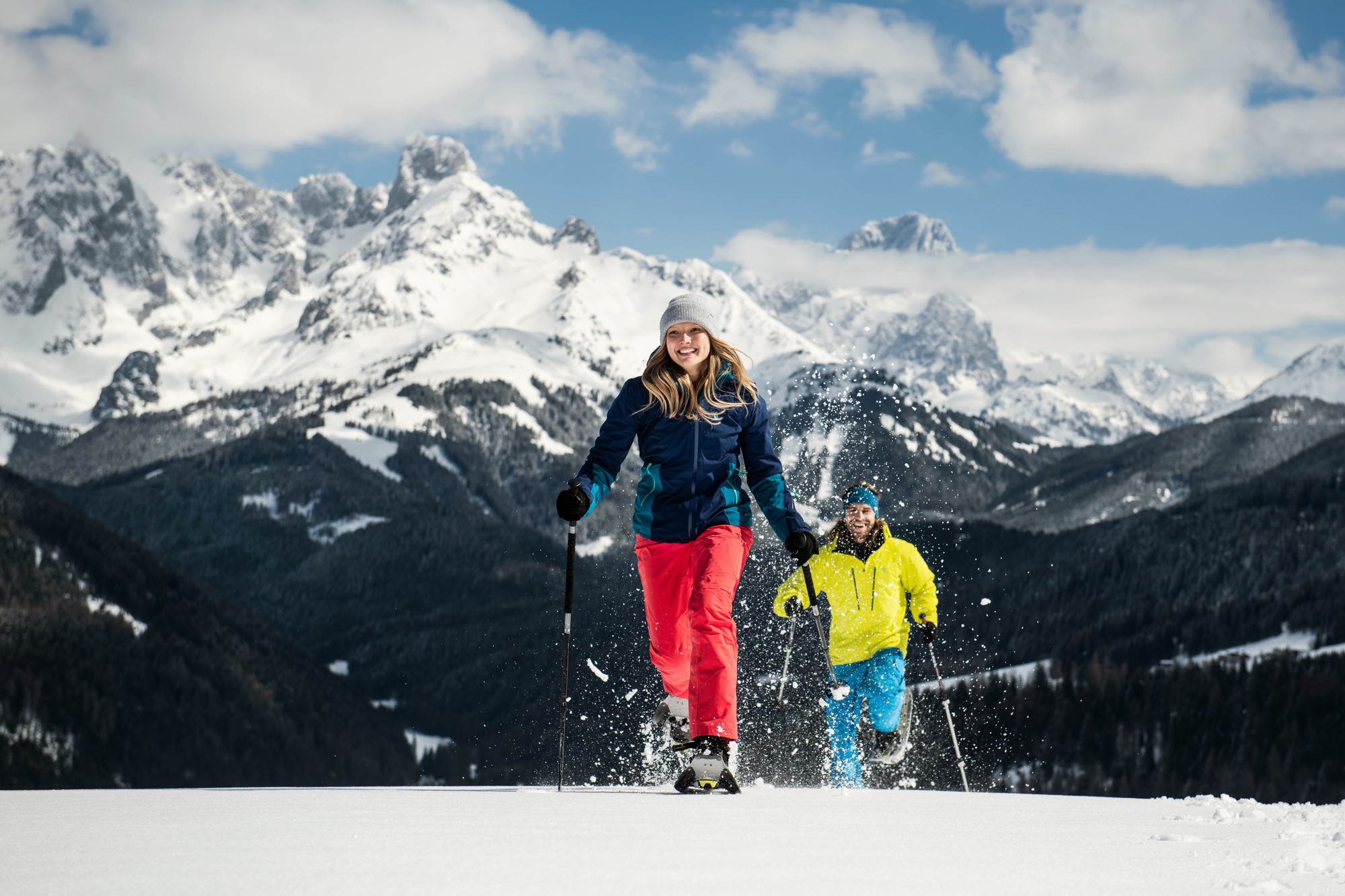 Zwei Personen beim Schneeschuhwandern auf einem verschneiten Berg mit malerischen Gipfeln und Wäldern im Hintergrund.