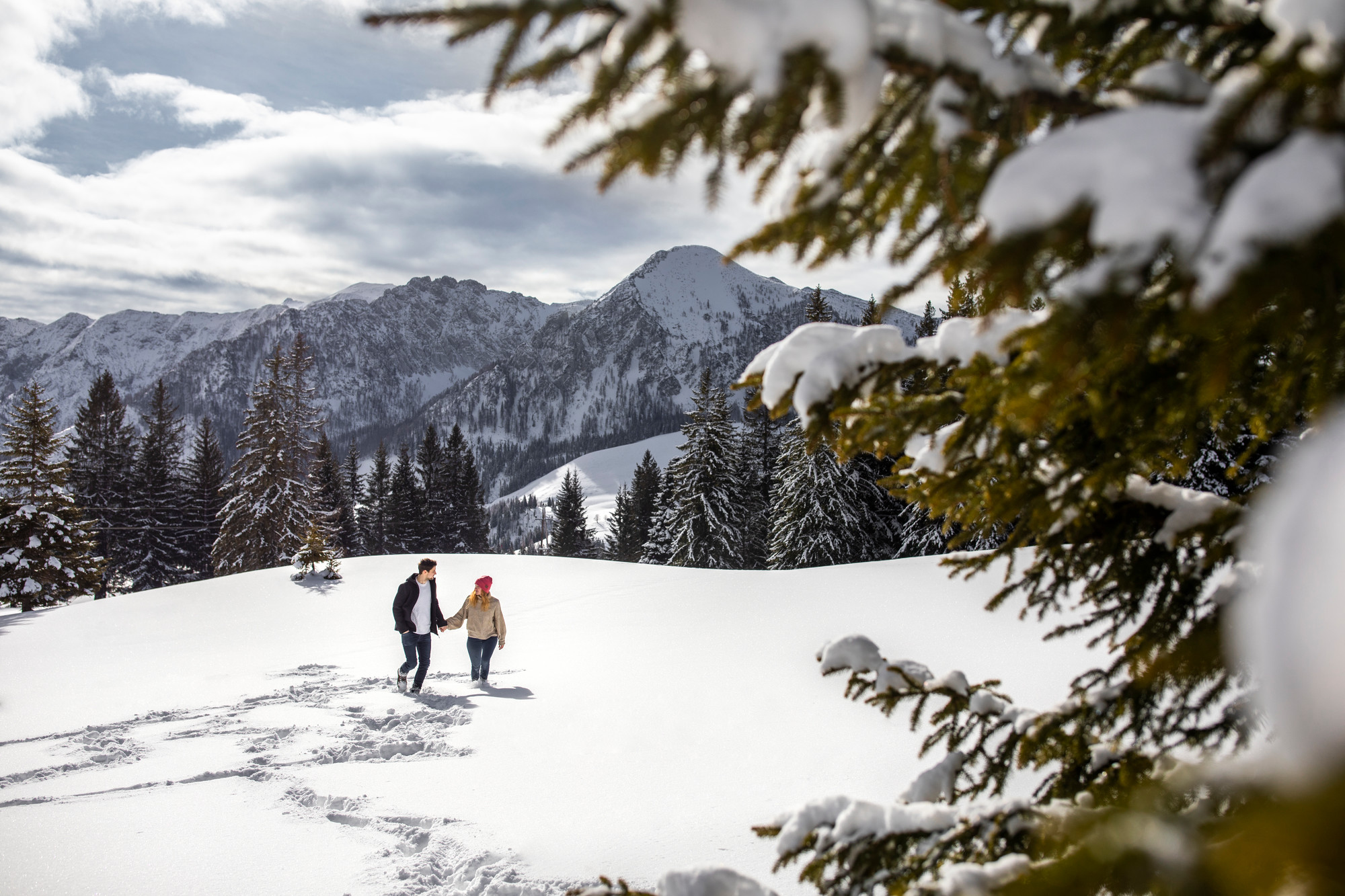 Ein Paar, das sich an den Händen hält und in den verschneiten Bergen spazieren geht, umgeben von Kiefern und schneebedeckter Landschaft.