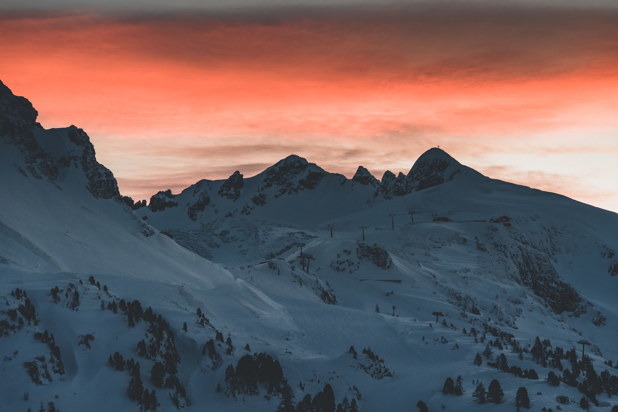 Verschneite Berggipfel mit Skiliften bei Sonnenuntergang unter einem dramatischen orange-grauen Himmel.