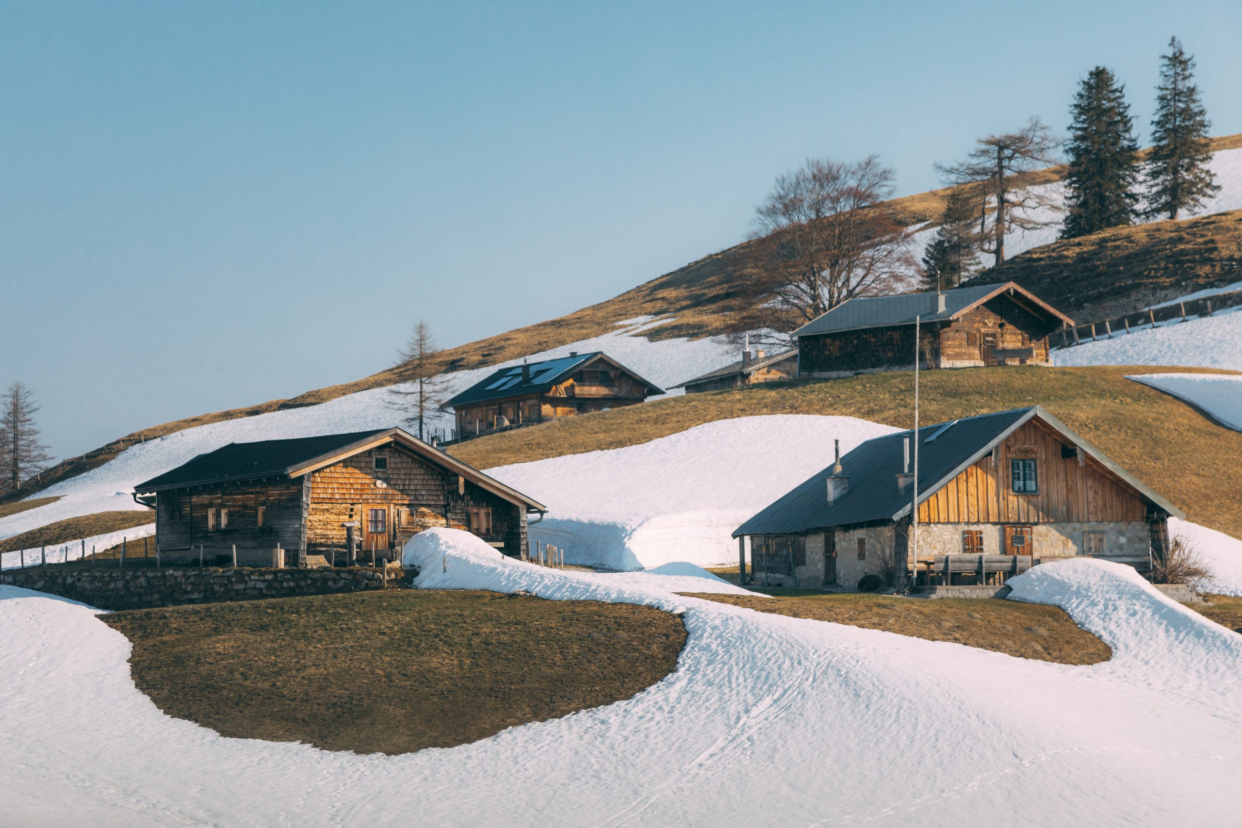 Holzhütten an einem verschneiten Hang mit Grasflächen und ein paar Bäumen unter einem klaren Himmel.