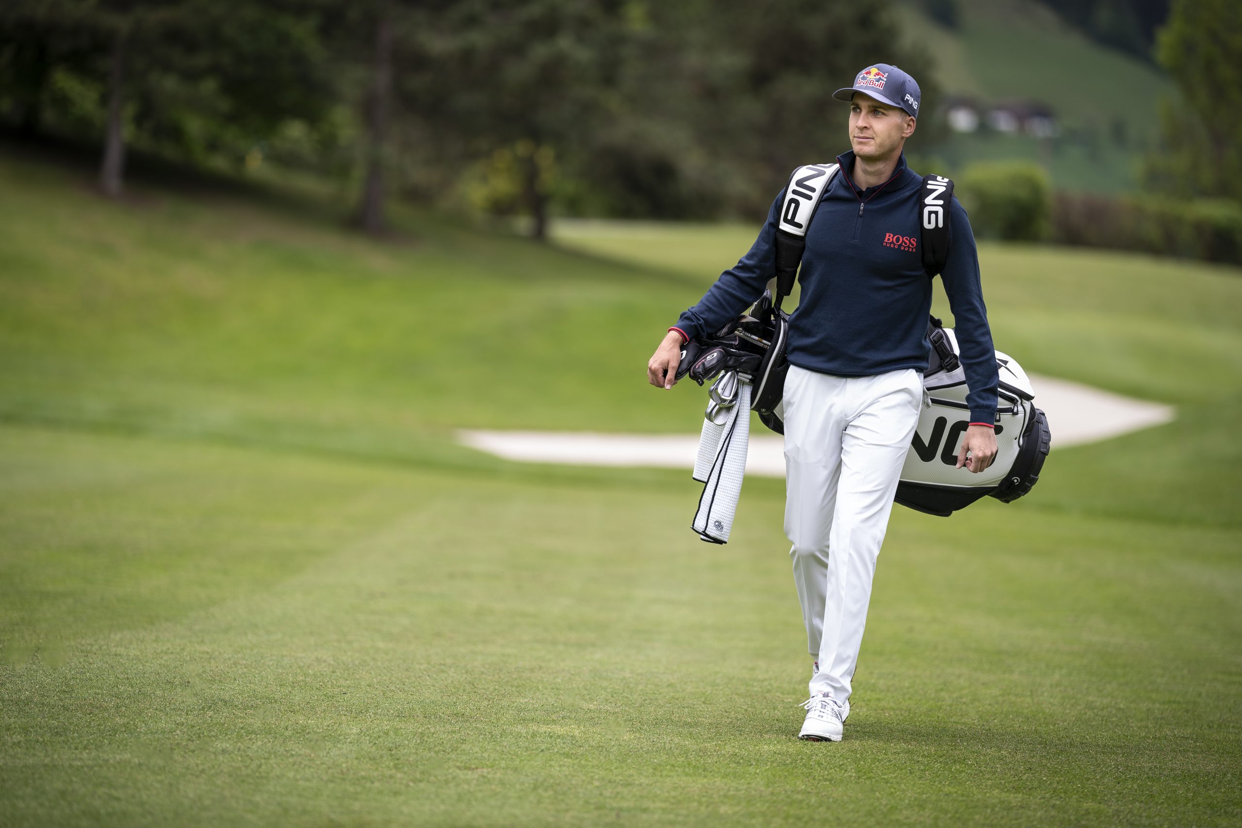 Ein Golfer mit einer Mütze geht auf einem Golfplatz und trägt eine Golftasche über der Schulter.