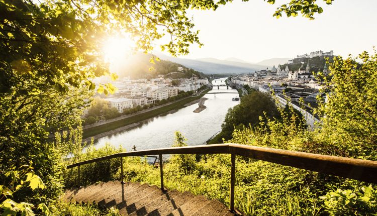 Eine geschwungene Steintreppe führt hinunter ins Grüne, mit Blick auf eine sonnenbeschienene Stadt am Fluss und die fernen Berge.