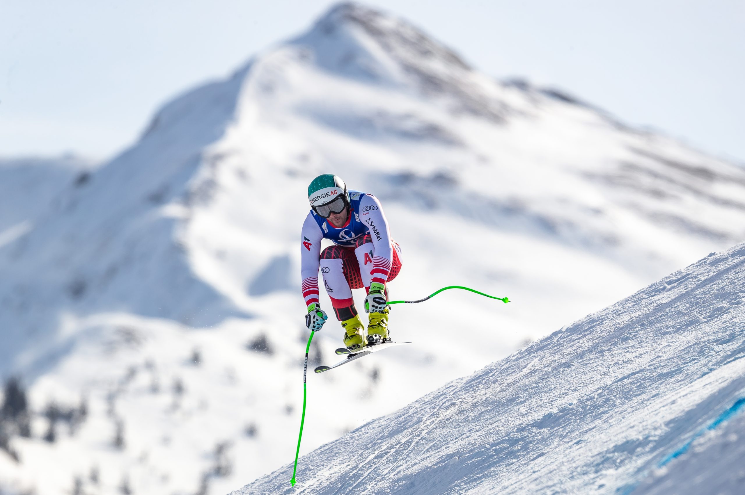 Ein Skifahrer in der Luft rast auf einem verschneiten Berg mit sichtbaren Gipfeln im Hintergrund bergab.