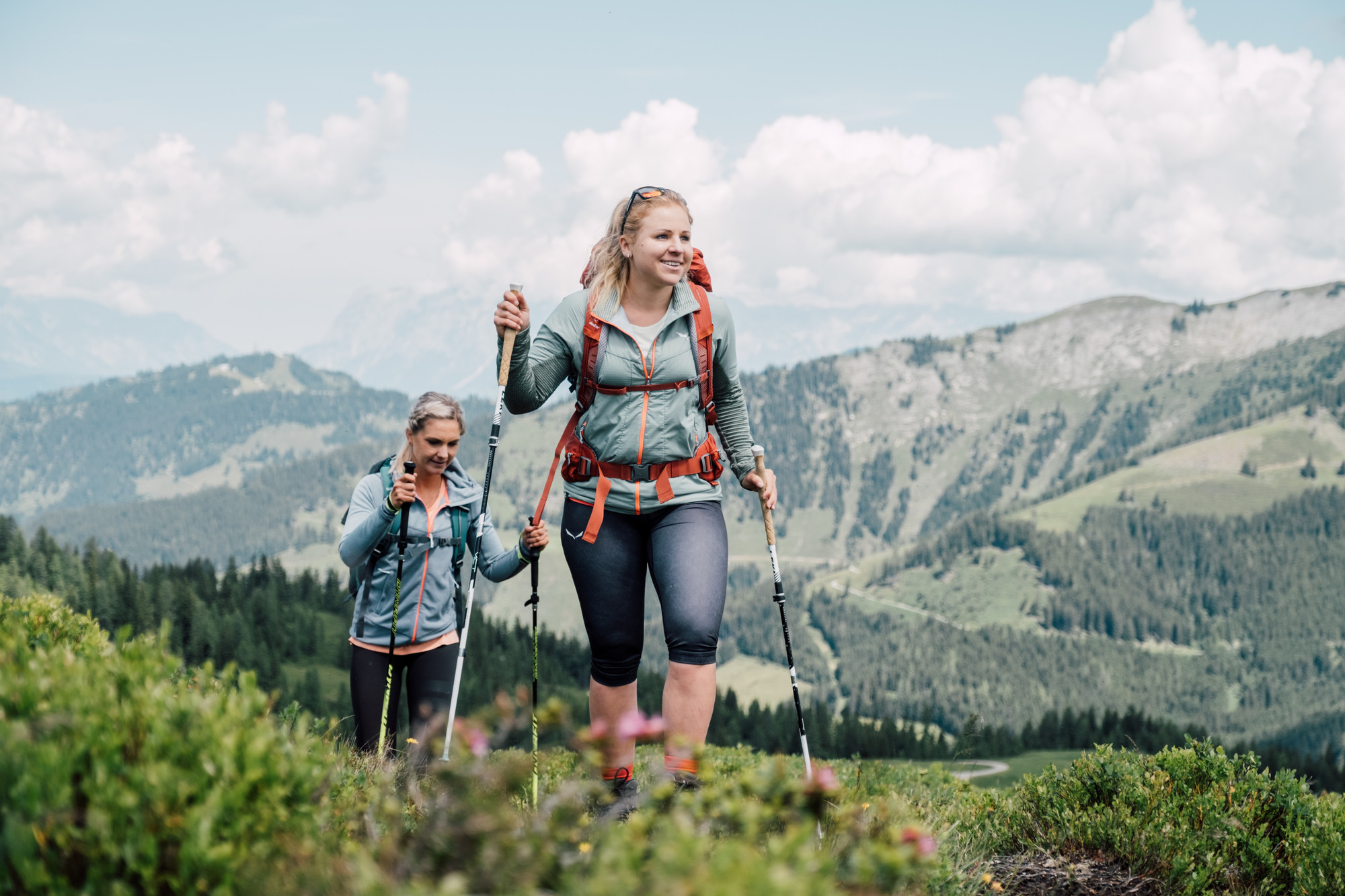 Zwei Frauen wandern mit Trekkingstöcken auf einem grünen Bergpfad mit malerischer Aussicht im Hintergrund.