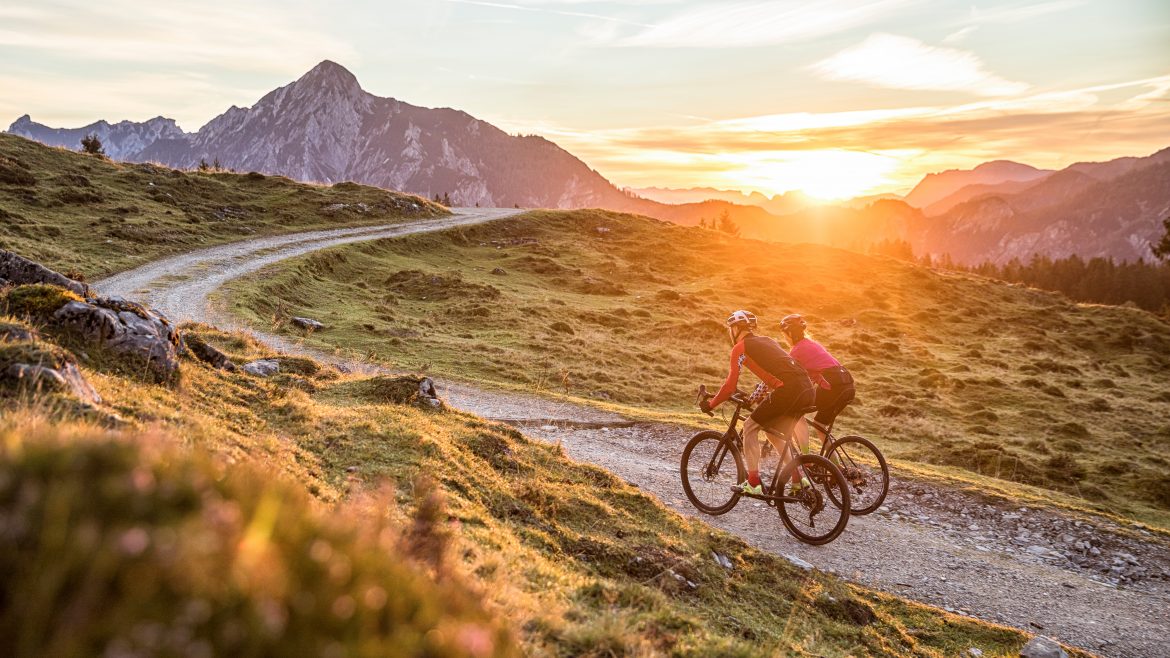 Zwei Radfahrer fahren auf einem Schotterweg durch grasbewachsene Hügel bei Sonnenuntergang, mit Bergen im Hintergrund.