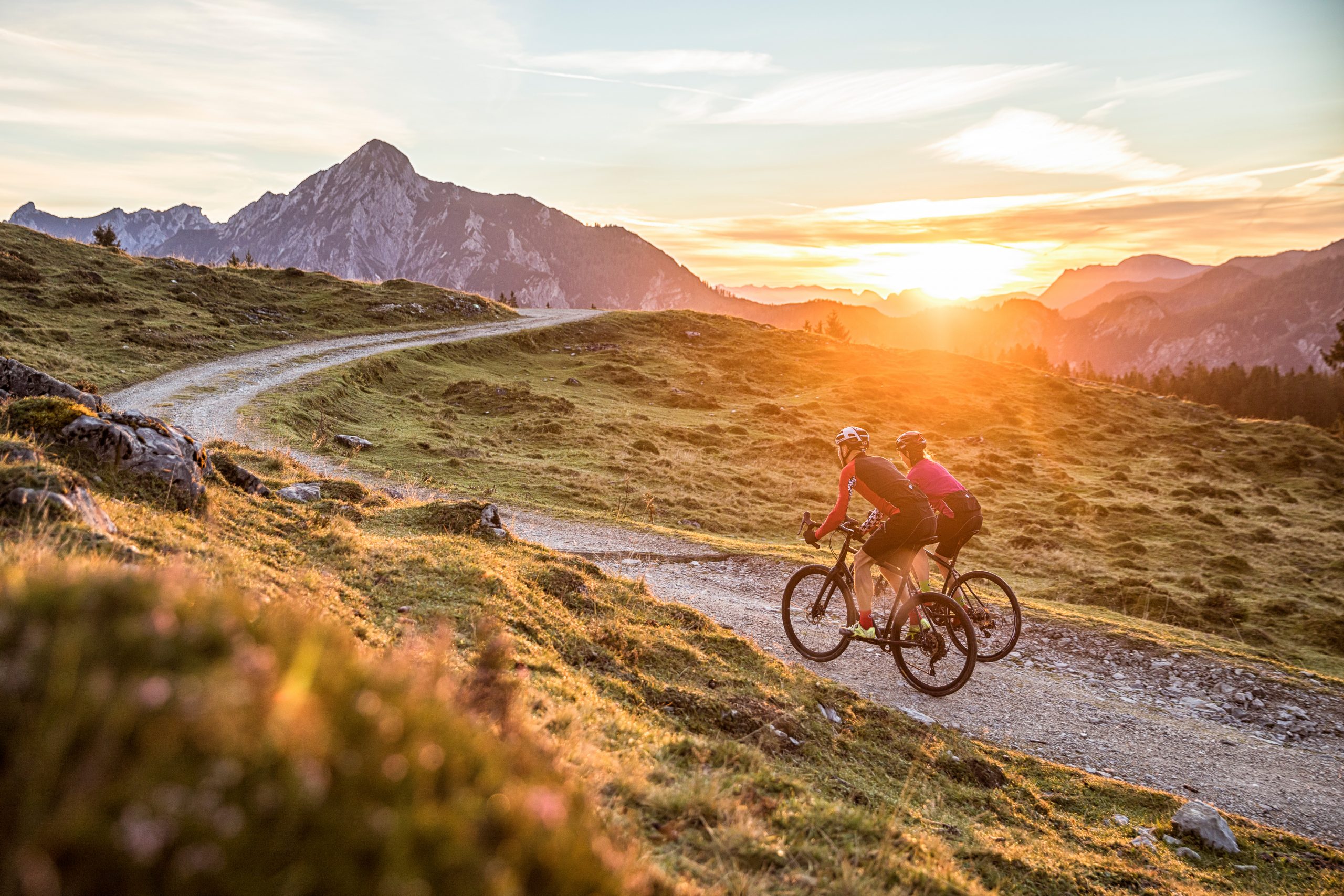 Zwei Radfahrer fahren auf einem Schotterweg durch grasbewachsene Hügel bei Sonnenuntergang, mit Bergen im Hintergrund.