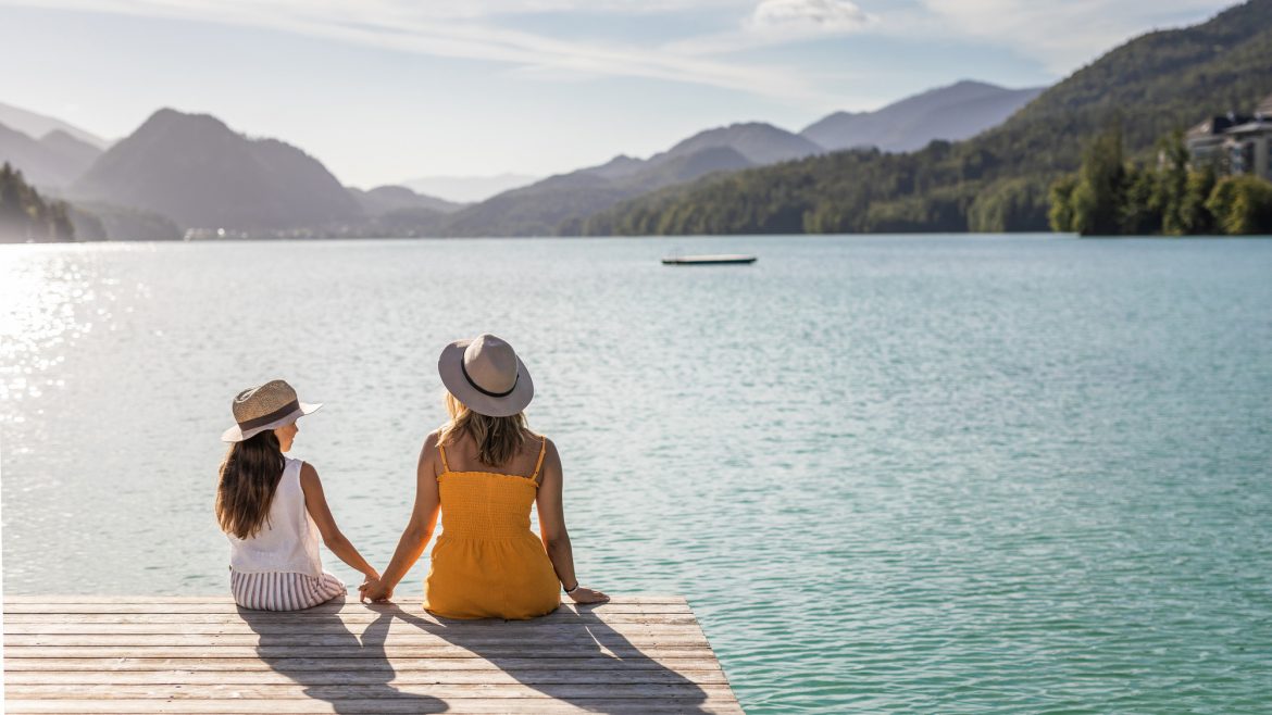 Zwei Personen mit Hüten sitzen an einem sonnigen Tag auf einem Steg mit Blick auf einen See und Berge.