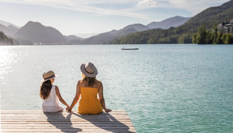 Zwei Personen mit Hüten sitzen an einem sonnigen Tag auf einem Steg mit Blick auf einen See und Berge.