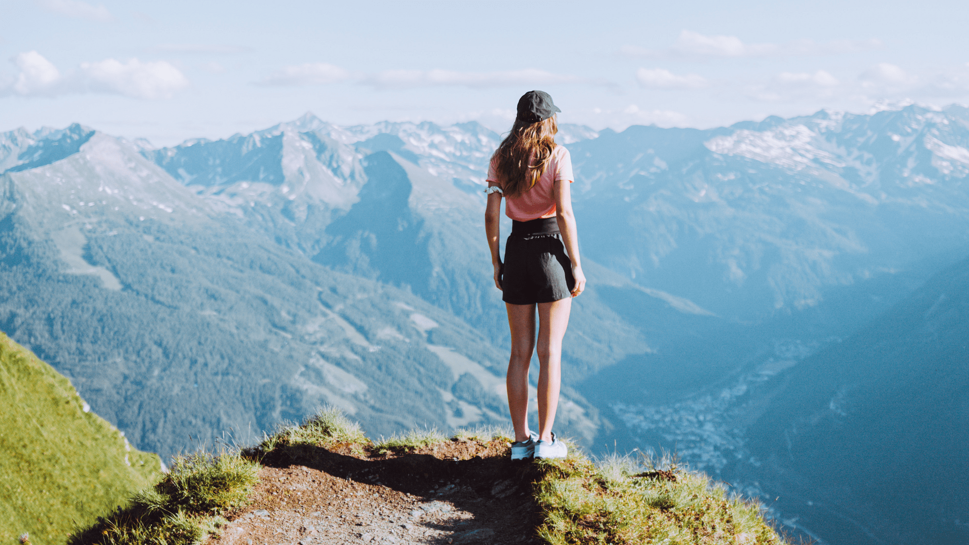 Eine Frau steht auf einer grasbewachsenen Klippe mit Blick auf die fernen Berge unter einem klaren blauen Himmel.