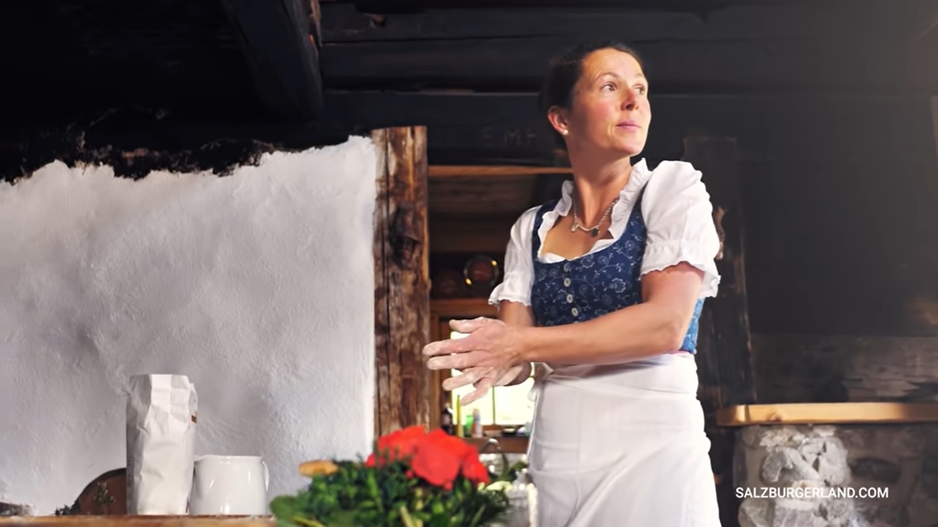 Eine Frau in traditioneller Kleidung steht in einer rustikalen Küche und wischt sich neben einer Vase mit roten Blumen das Mehl von den Händen.