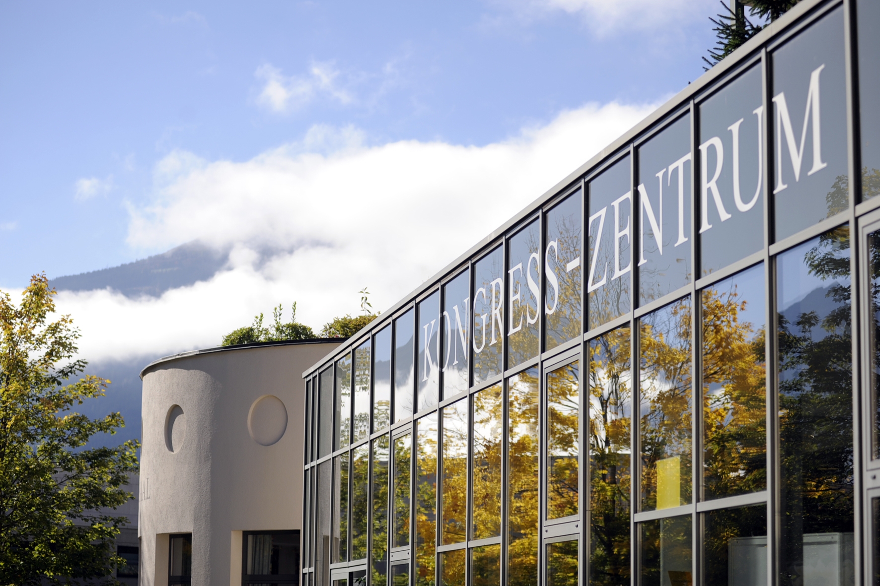 Modernes Glasgebäude mit der Aufschrift Kongress-Zentrum mit Bäumen und Bergen im Hintergrund.